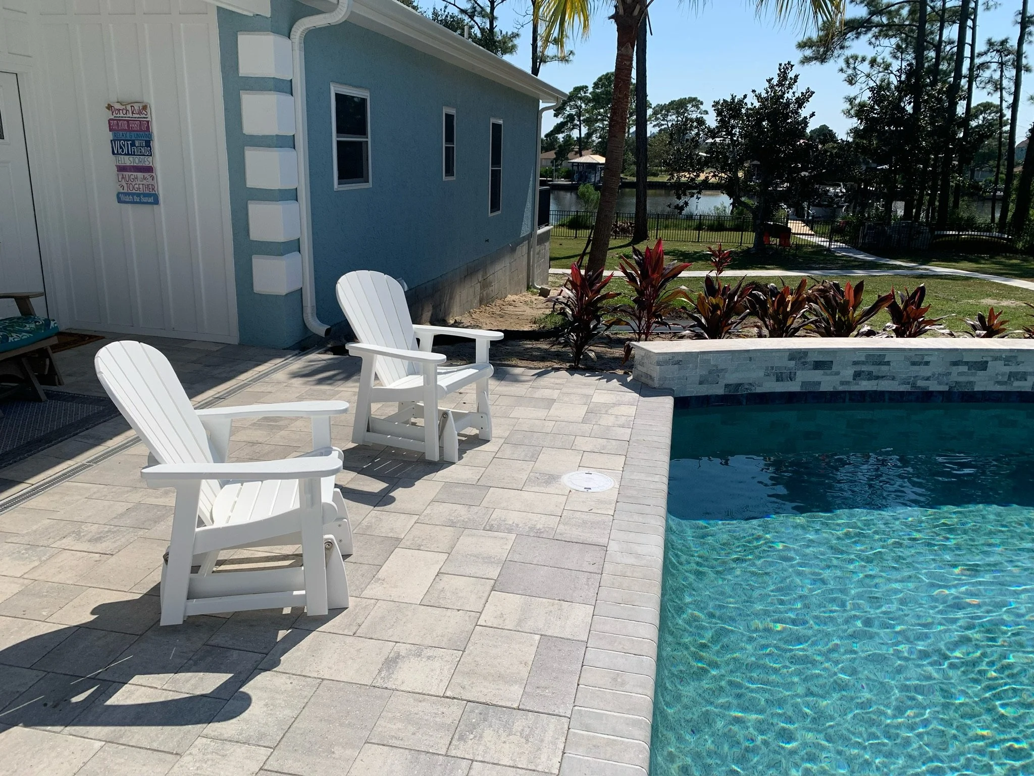A backyard scene with a swimming pool in the foreground, surrounded by a tiled patio area with white Adirondack chairs. There is a house with a blue exterior wall and windows, a sign with decorative writing on the outdoor wall, and landscaped bushes 