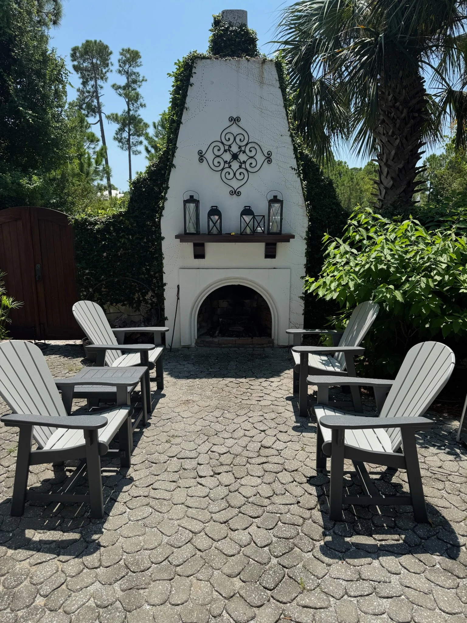 An outdoor patio with six white Adirondack chairs arranged in pairs facing a white fireplace with a decorative iron wall hanging and three lanterns on a wooden shelf. The patio is paved with cobblestone, surrounded by greenery including trees and bus