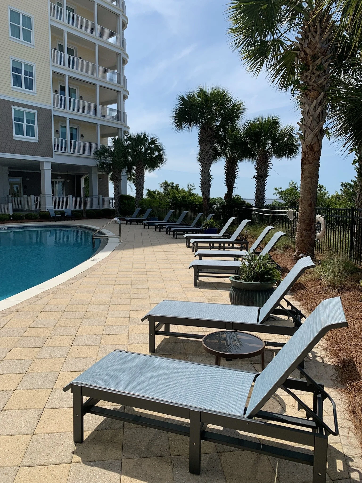 Swimming pool area at a resort with lounge chairs, palm trees, and a multi-story building in the background.
