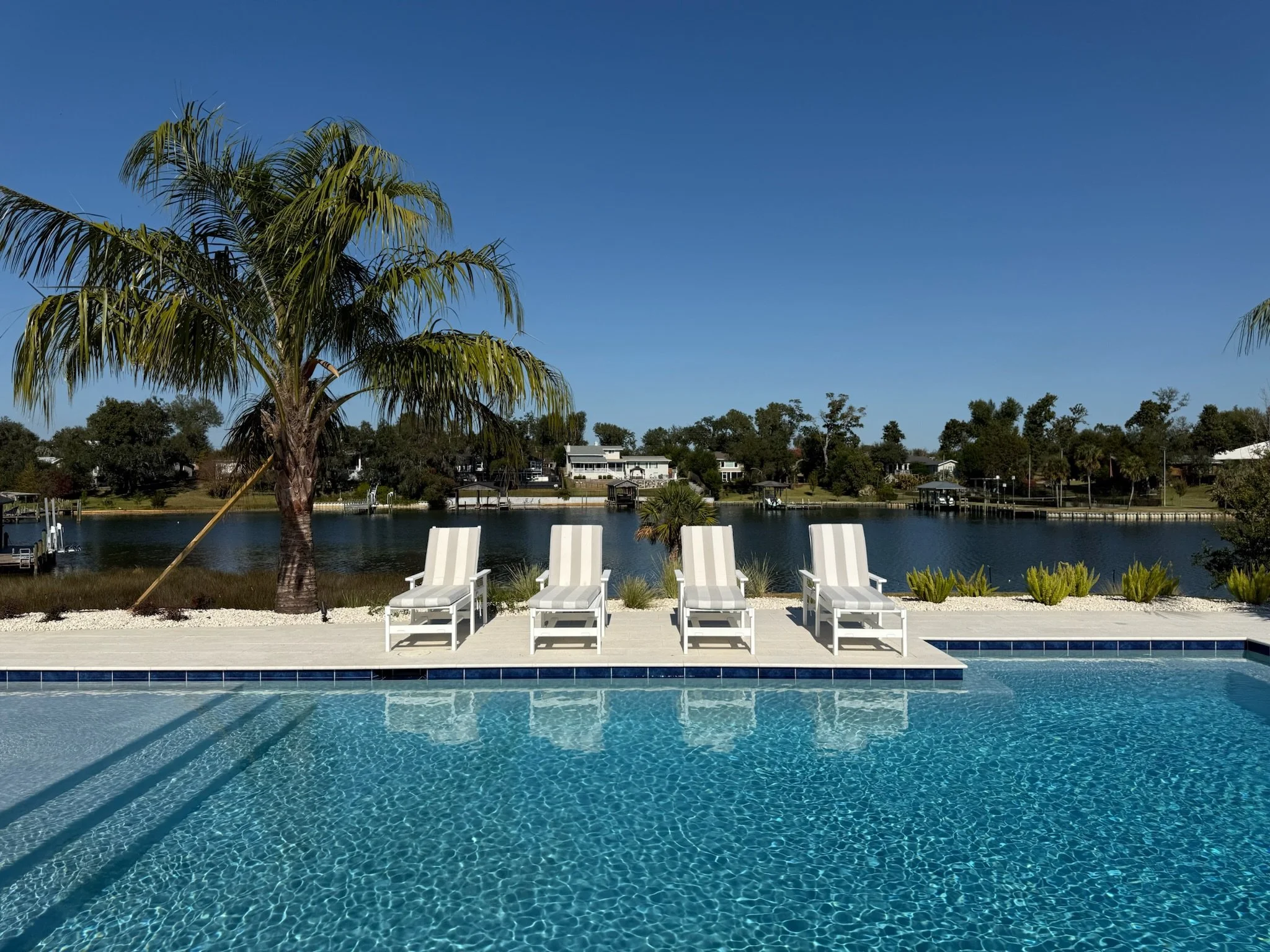 A backyard pool area with four white lounge chairs on a tiled deck, overlooking a body of water with houses and trees in the background and a clear blue sky.