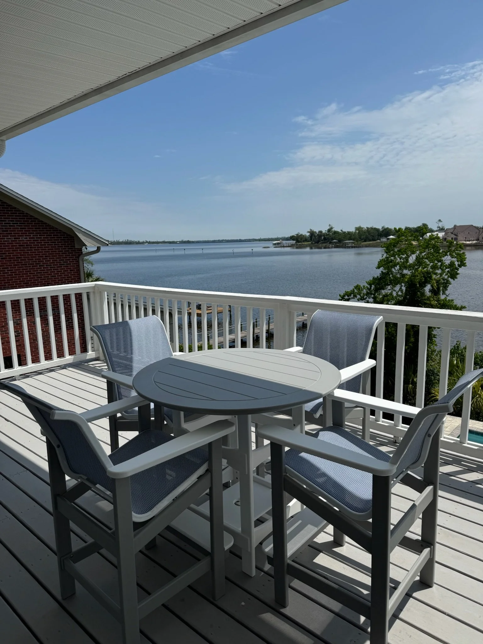 A white outdoor patio table with six chairs on a balcony overlooking a body of water with a cloudy sky and distant houses.