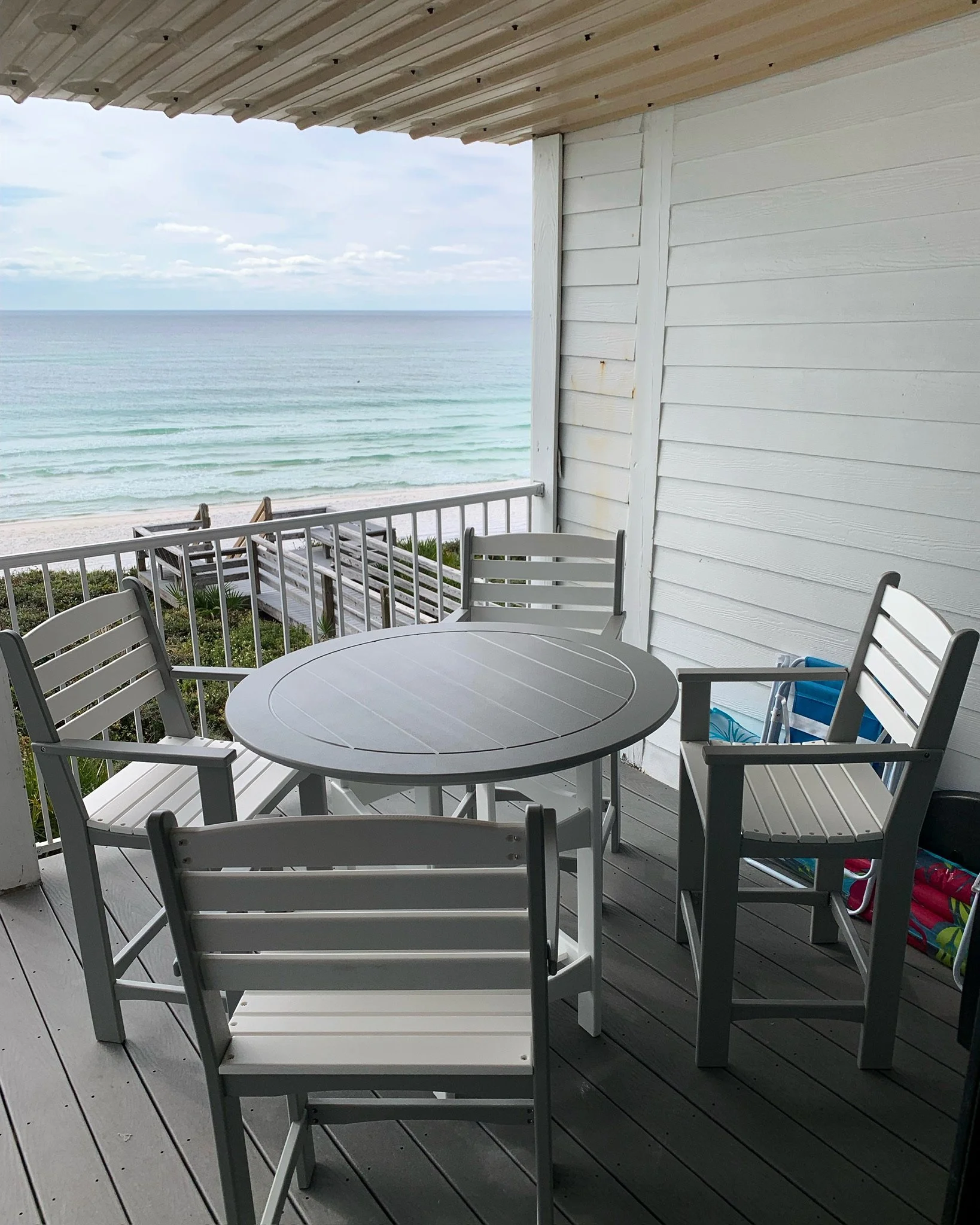 View from a porch overlooking a beach and ocean, with white wooden furniture including a round table and several chairs, and other patio items visible.