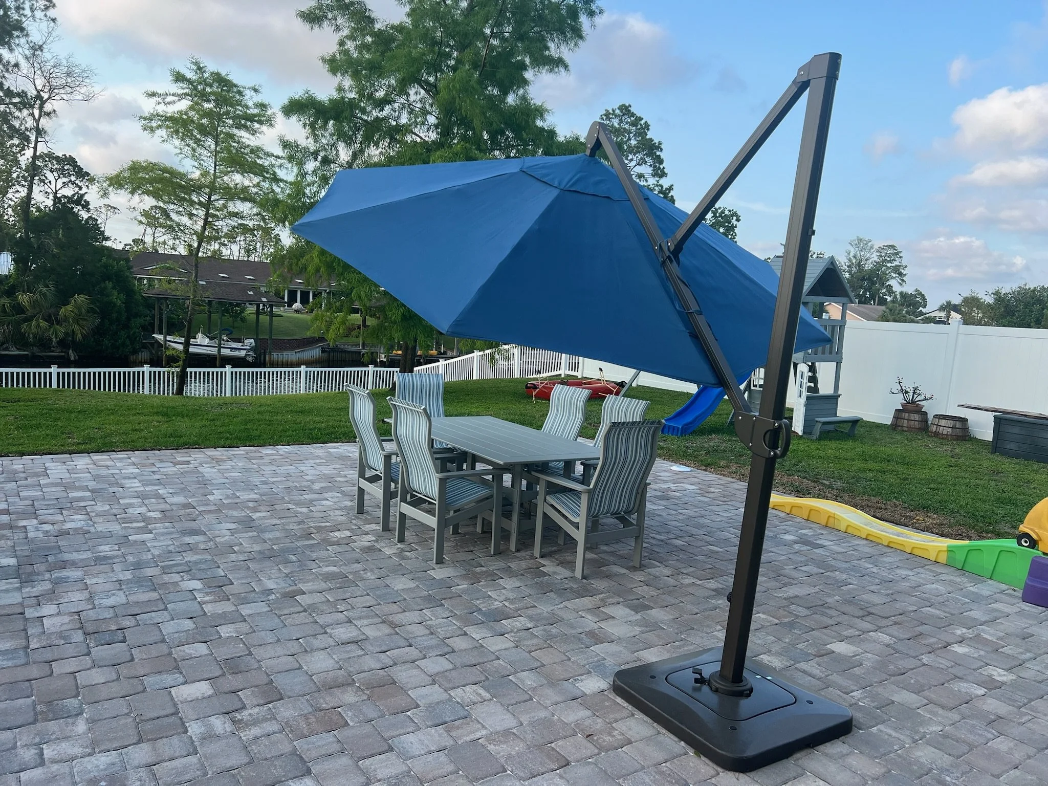 Backyard patio with a large blue patio umbrella, a gray outdoor dining table with six striped chairs, a small slide, a yellow toy car, and a white fence surrounding the yard. Trees and houses are visible in the background.
