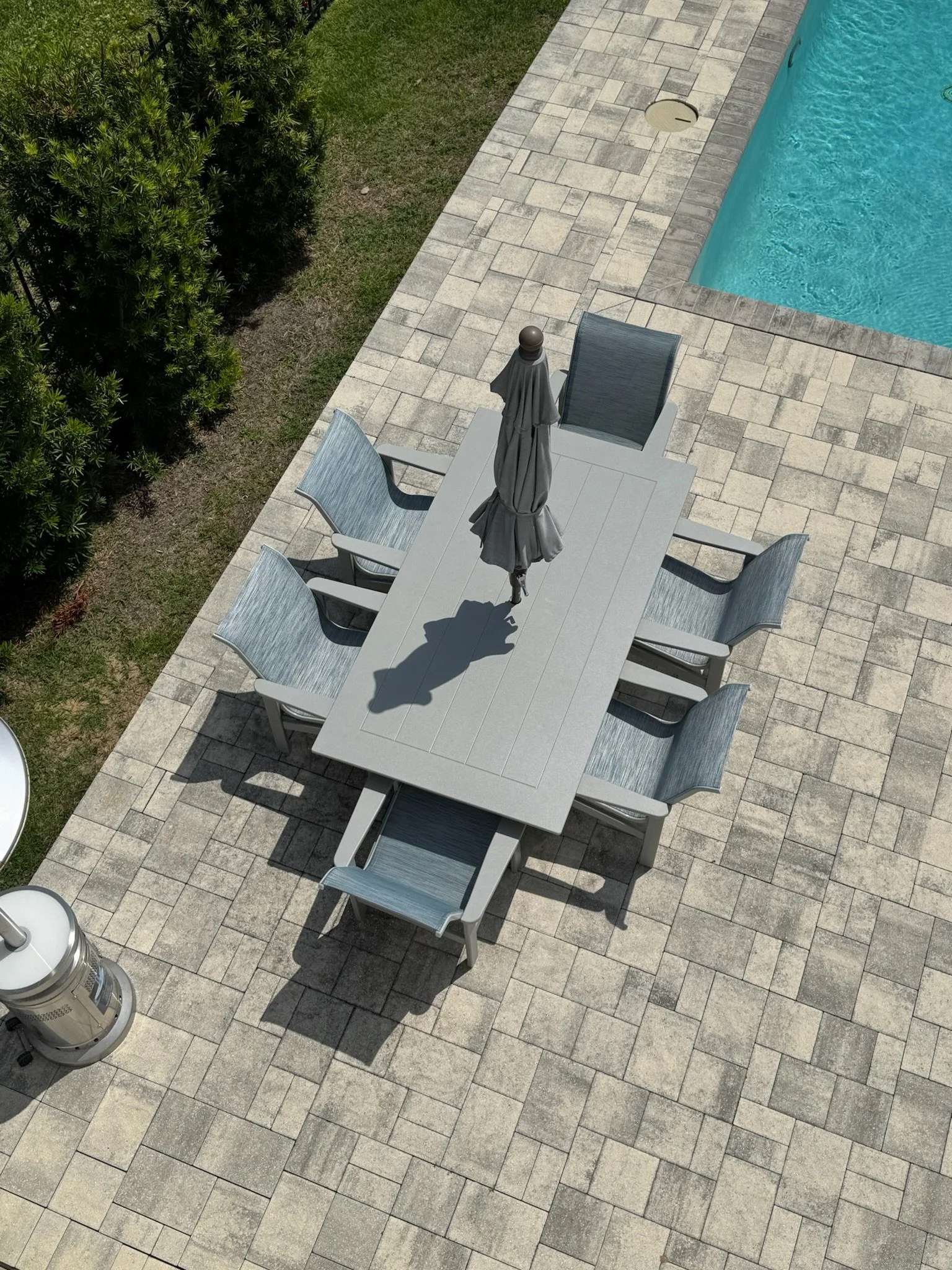 Overhead view of an outdoor patio with a large rectangular table, six chairs, a closed umbrella, a swimming pool, and a gas heater, with a grassy area and bushes nearby.