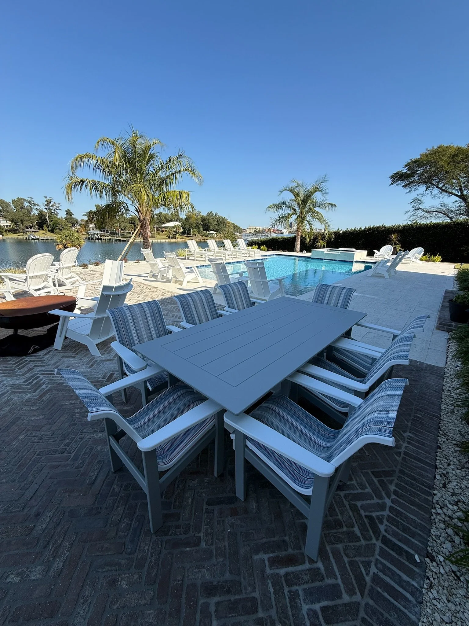 Outdoor pool area with white lounge chairs, umbrellas, and a view of a waterway with houses, trees, and a clear blue sky.