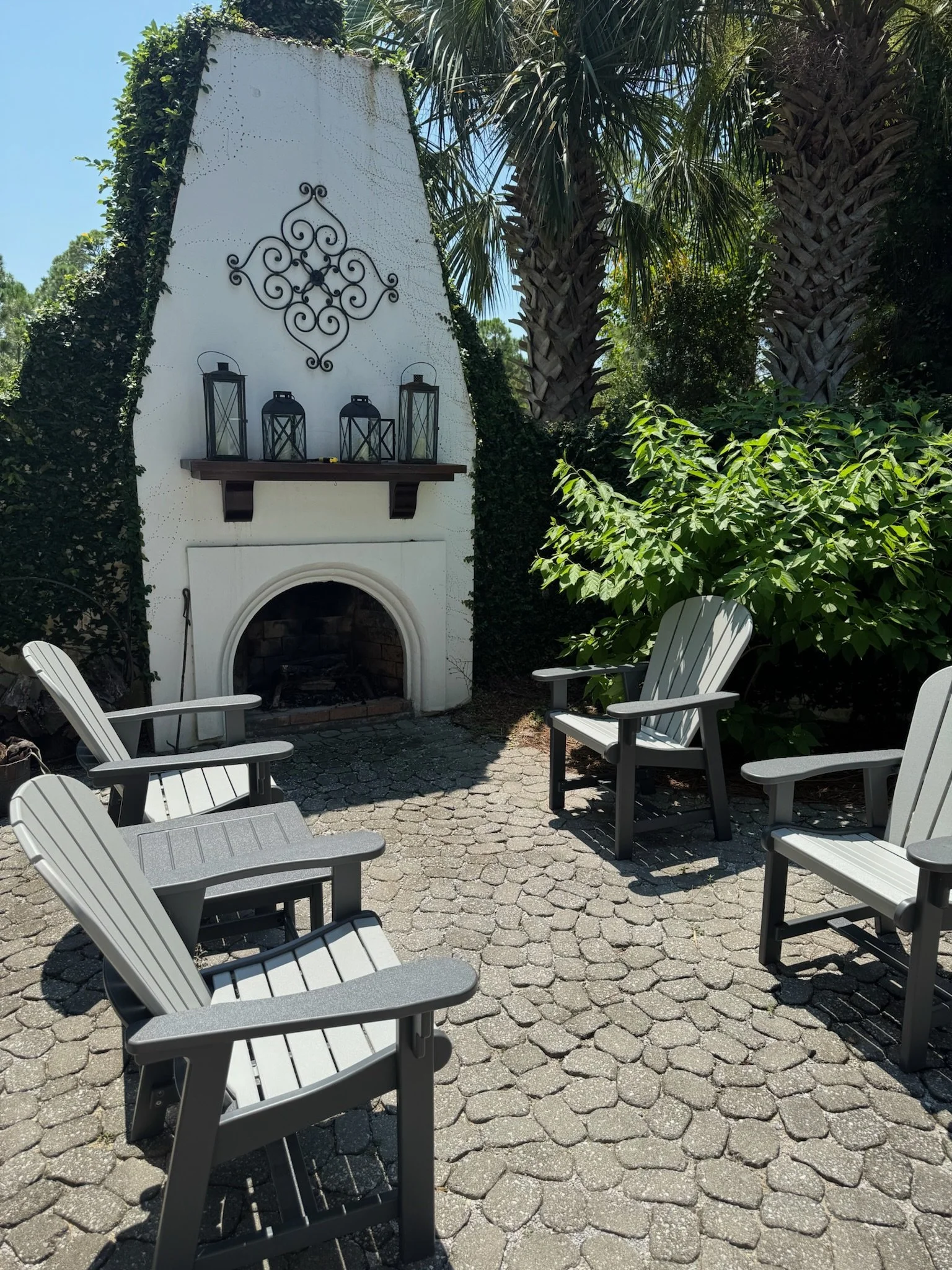 An outdoor seating area with gray chairs arranged on a cobblestone patio, adjacent to a white chimney with a fire opening. The chimney is decorated with black wrought iron scrollwork and surrounded by lush green trees and plants, including palm trees