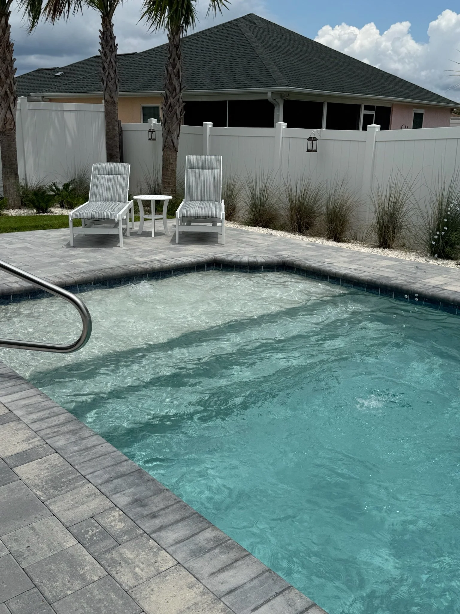 A backyard pool area with two white lounge chairs with striped cushions and a small white table between them, surrounded by a gray stone deck. Behind the lounge chairs, there are three tall palm trees, a white privacy fence, and some decorative plant
