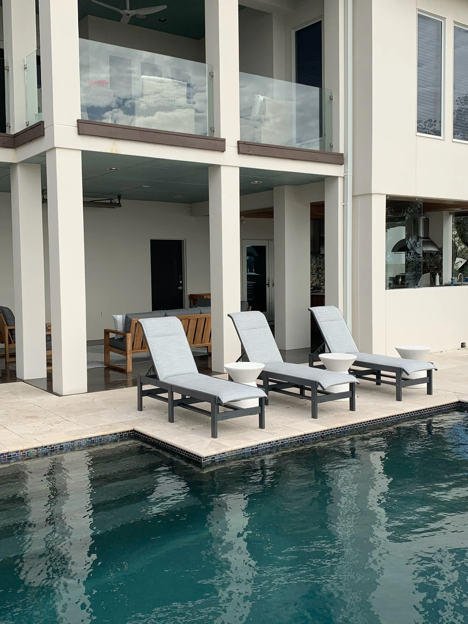 View of a backyard with a swimming pool in front, surrounded by a beige tiled patio area, with lounge chairs and small tables near the pool, and a modern house with large windows and a balcony in the background.