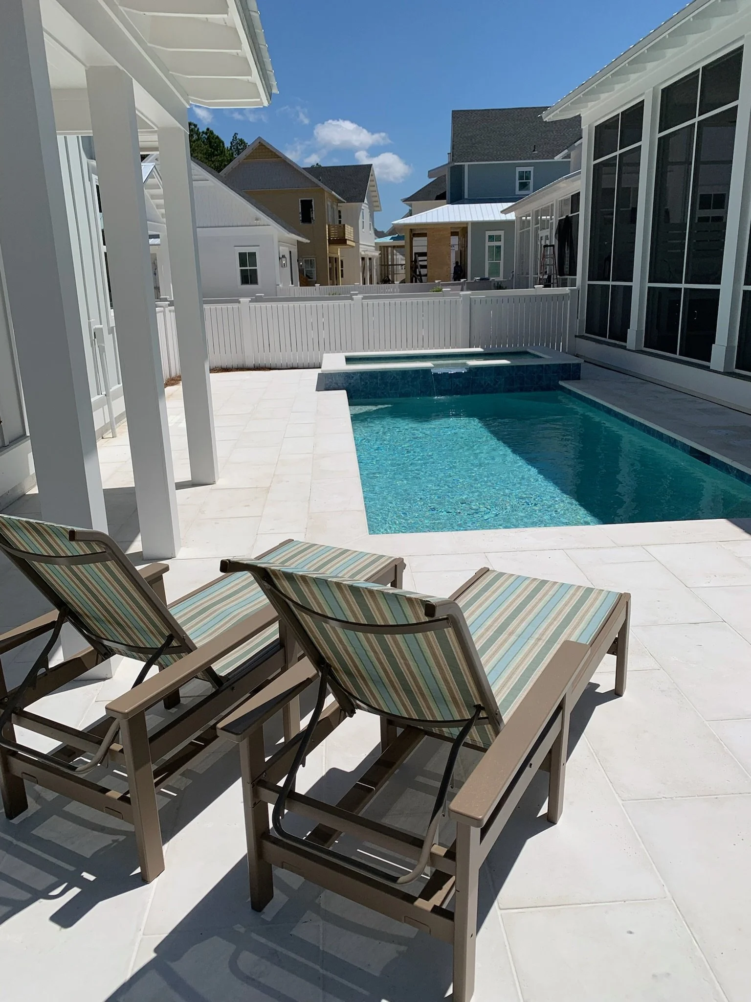 Backyard with a swimming pool, white fence, sun loungers with striped cushions, and neighboring houses under a bright blue sky.