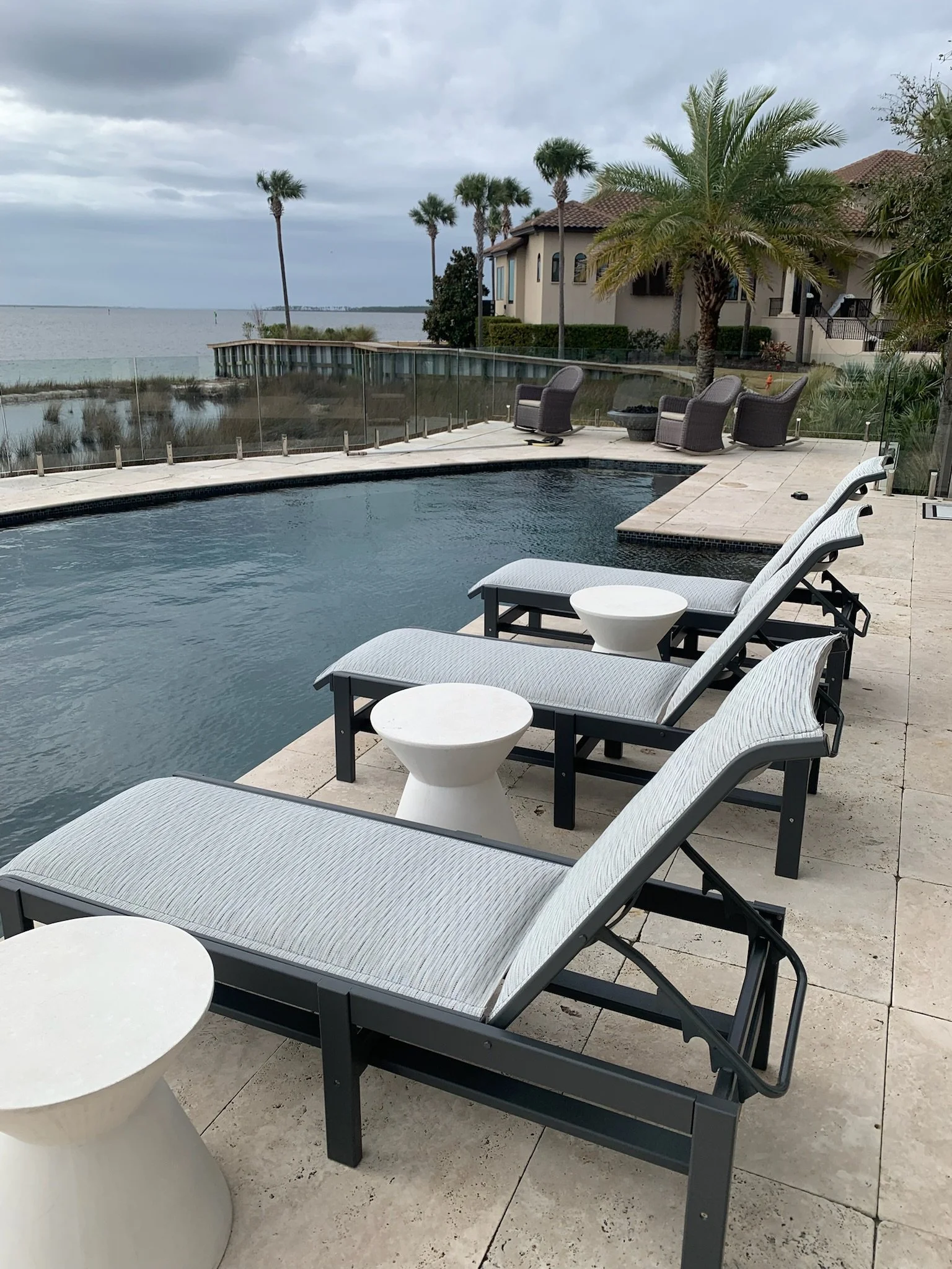 Poolside area with lounge chairs and small tables, overlooking a harbor with palm trees and houses under a cloudy sky.