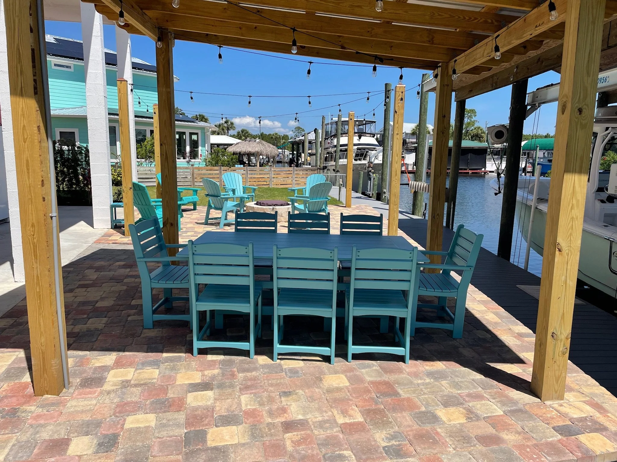 View of a backyard patio area with blue wooden chairs around a fire pit, under a wooden pergola with string lights, overlooking a waterway with boats and a house with a teal exterior in the background.