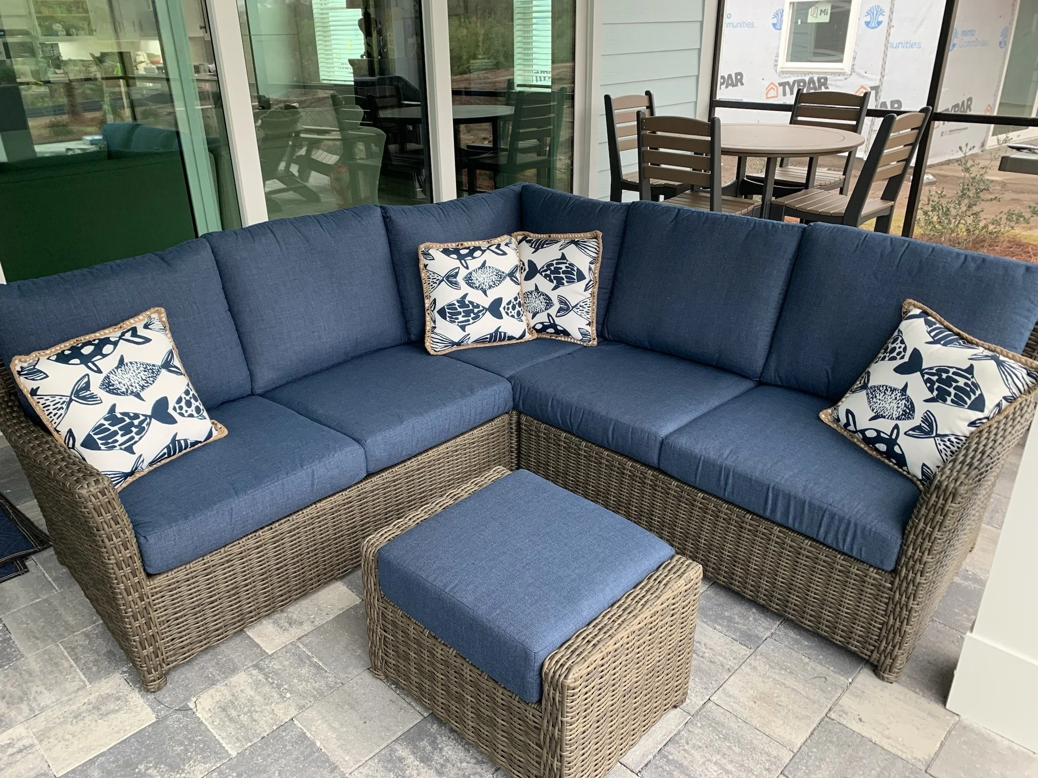 Outdoor patio furniture including a wicker sofa with blue cushions and patterned pillows, an ottoman, and a dining table with six chairs, next to a house with large glass windows and a sliding door, under construction with siding partially installed.