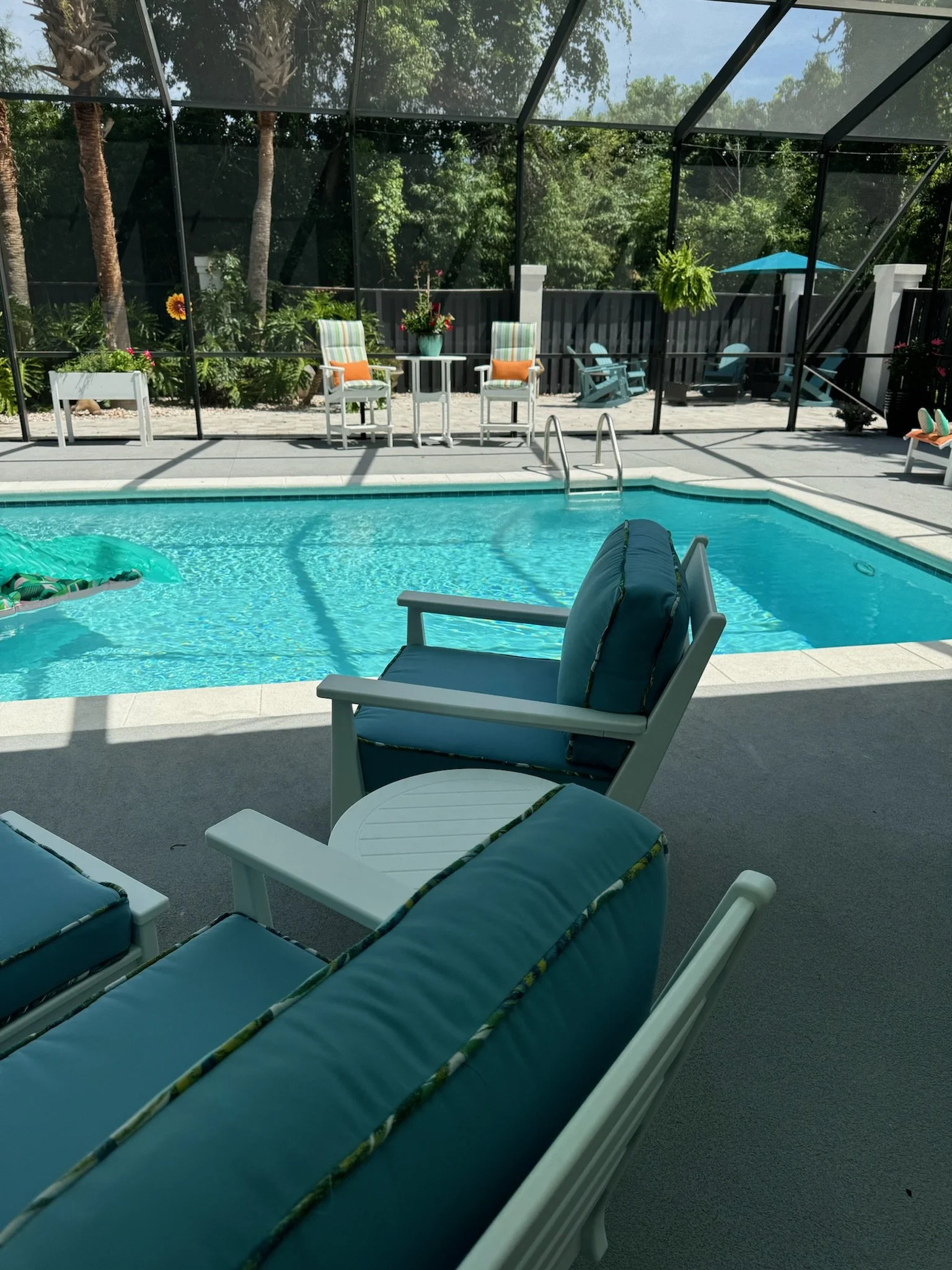 View of a backyard swimming pool area, enclosed with a screened structure. There are blue cushioned lounge chairs, patio chairs, plants, and a small table with an orange pillow. A blue umbrella and a shaded area with outdoor seating and potted plants