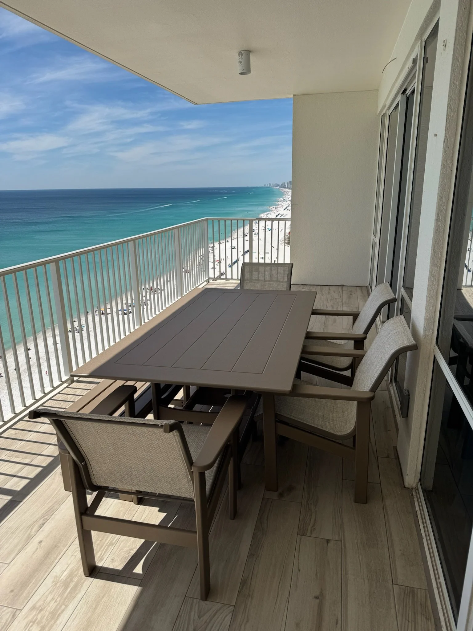 Balcony with a gray table and six chairs, overlooking a beach and ocean with a city skyline in the distance under a partly cloudy blue sky.