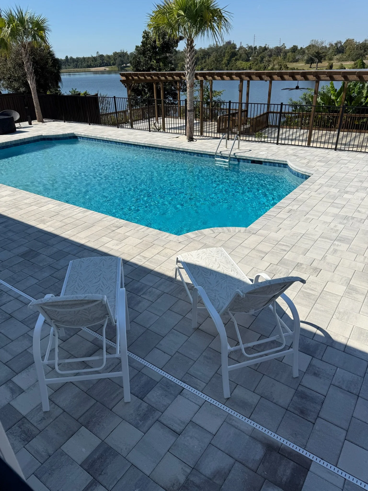 Swimming pool with white tiled deck, surrounded by palm trees, overlooking a lake with trees in the background, blue sky, and a pergola with stairs leading down to the water.