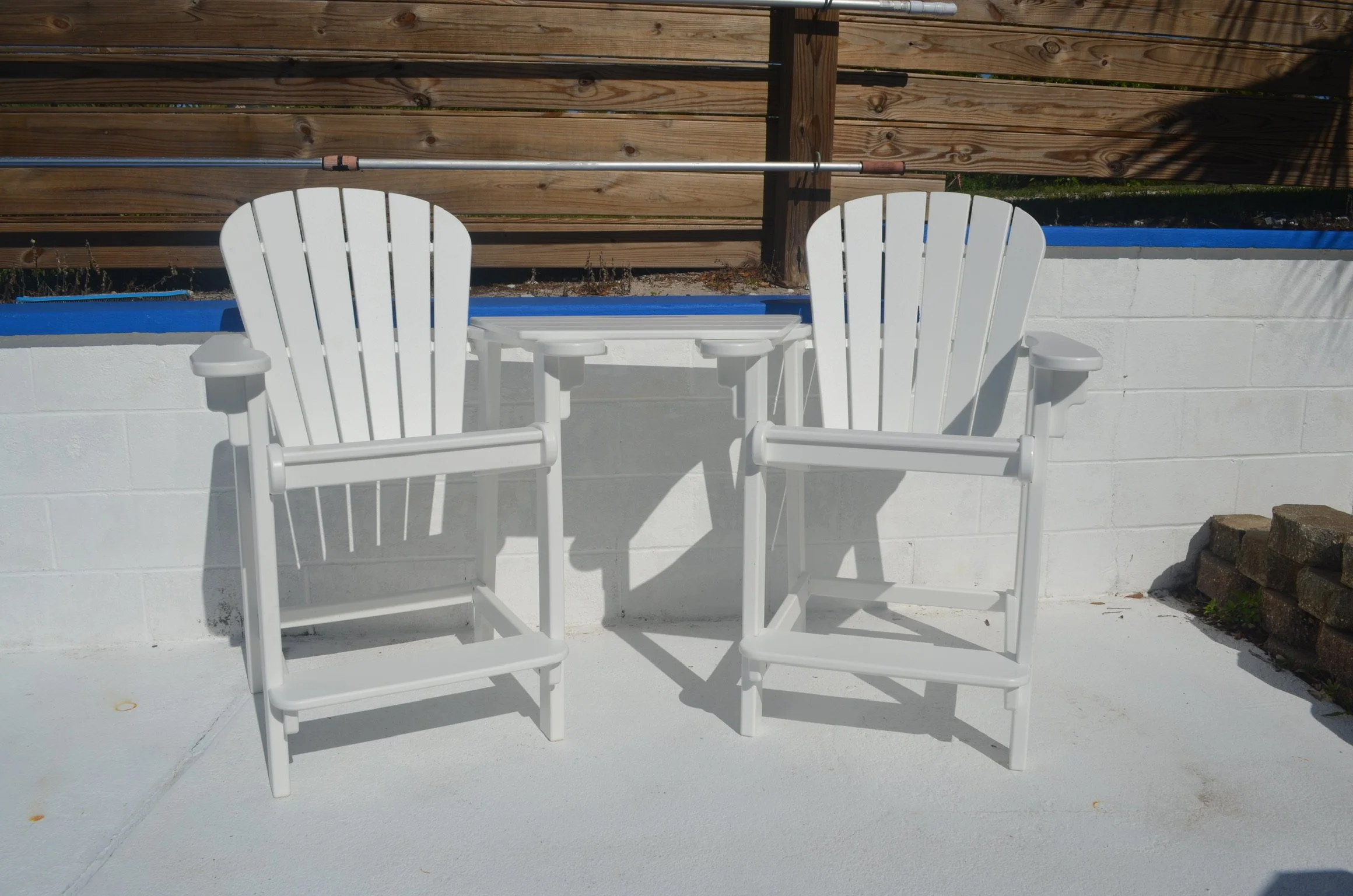 Two white Adirondack chairs on a white concrete patio with a wooden privacy fence and a gardening hand tool laying across the backrest of one chair.