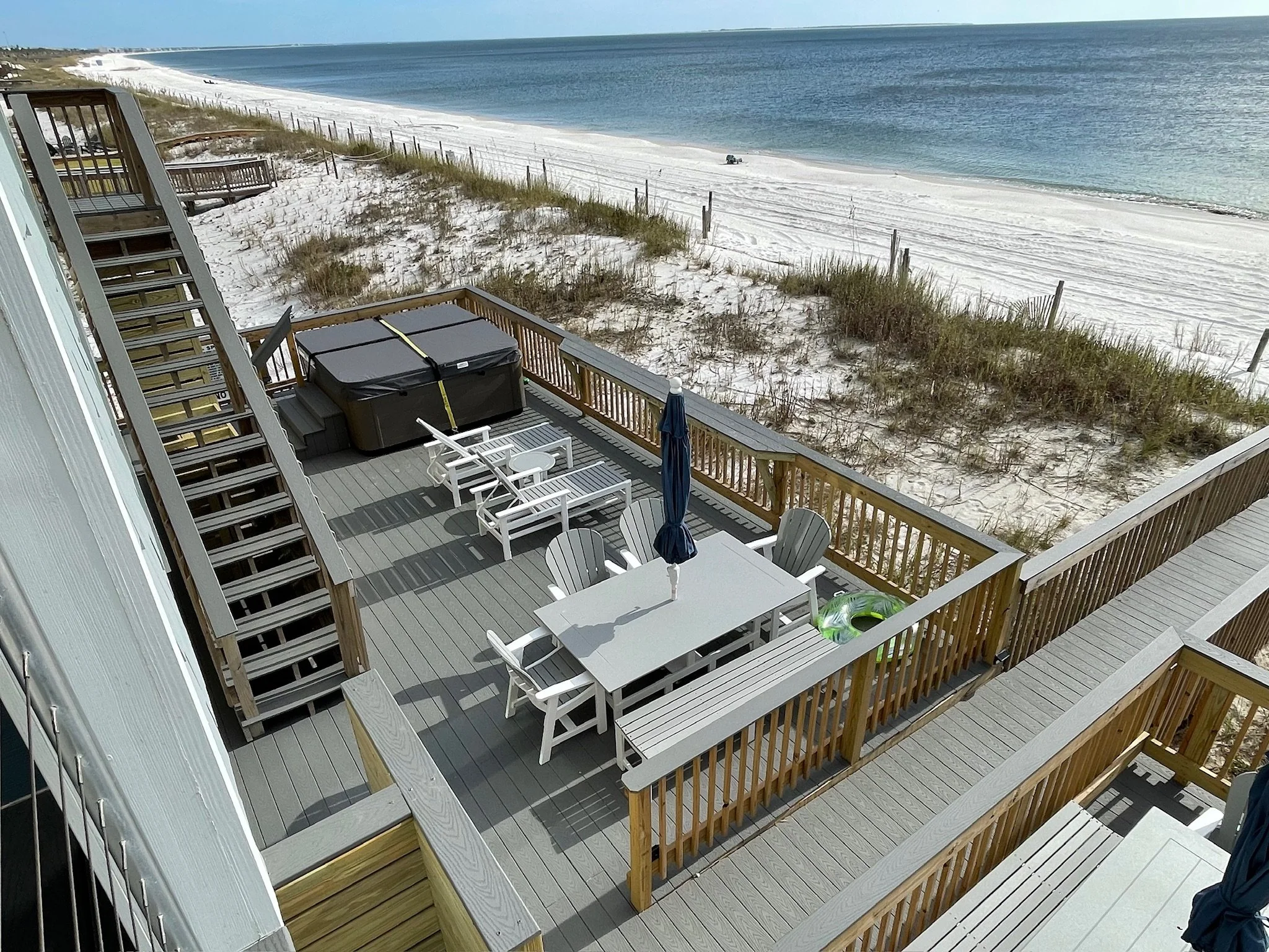 View from a balcony overlooking a sandy beach with white sand, dunes with grass, and ocean water under a clear blue sky.