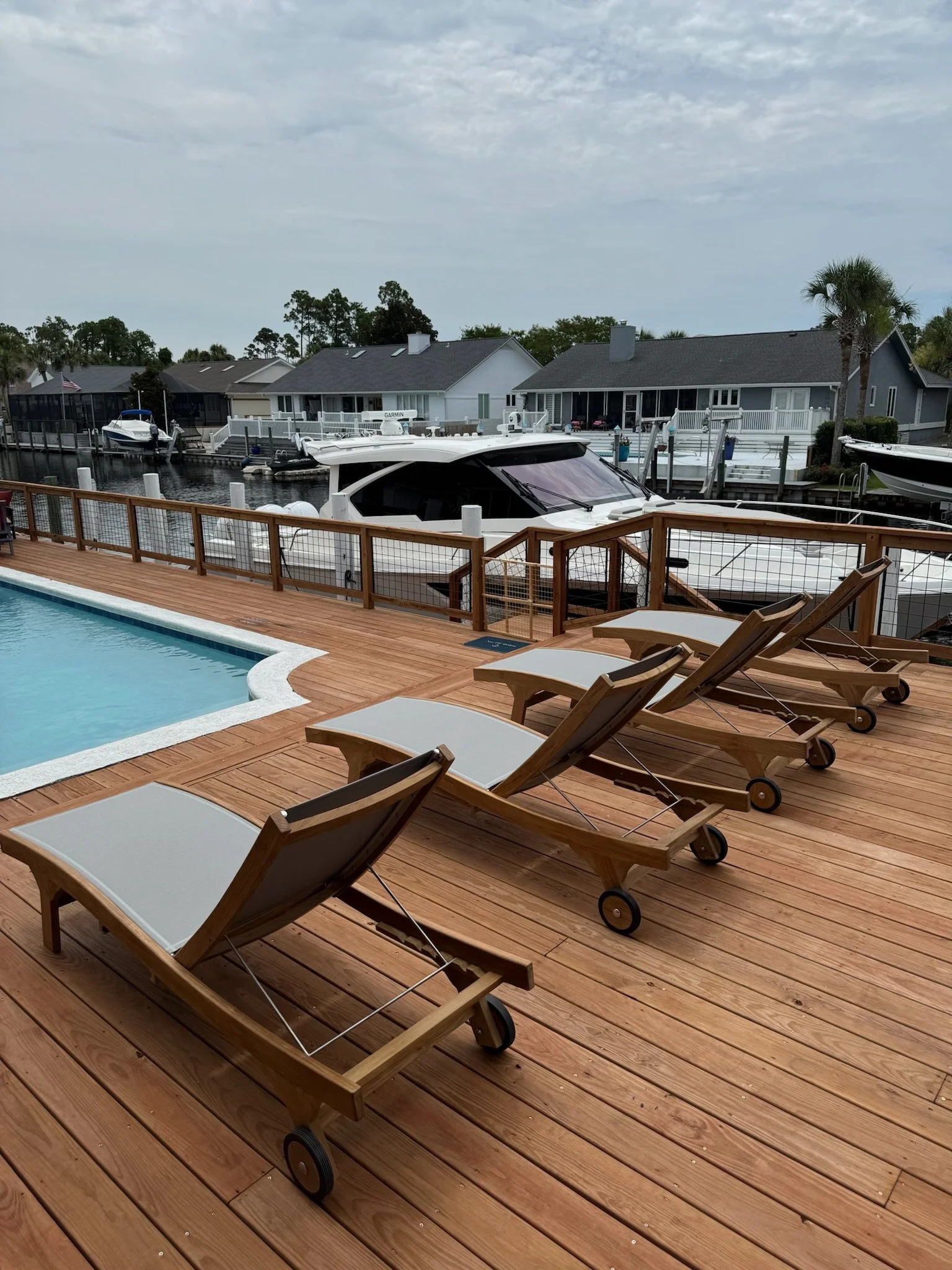 A backyard with a swimming pool and wooden deck, featuring four wooden lounge chairs with white cushions. In the background, there are houses, a boat docked at a waterway, and boats on the water, with cloudy skies overhead.