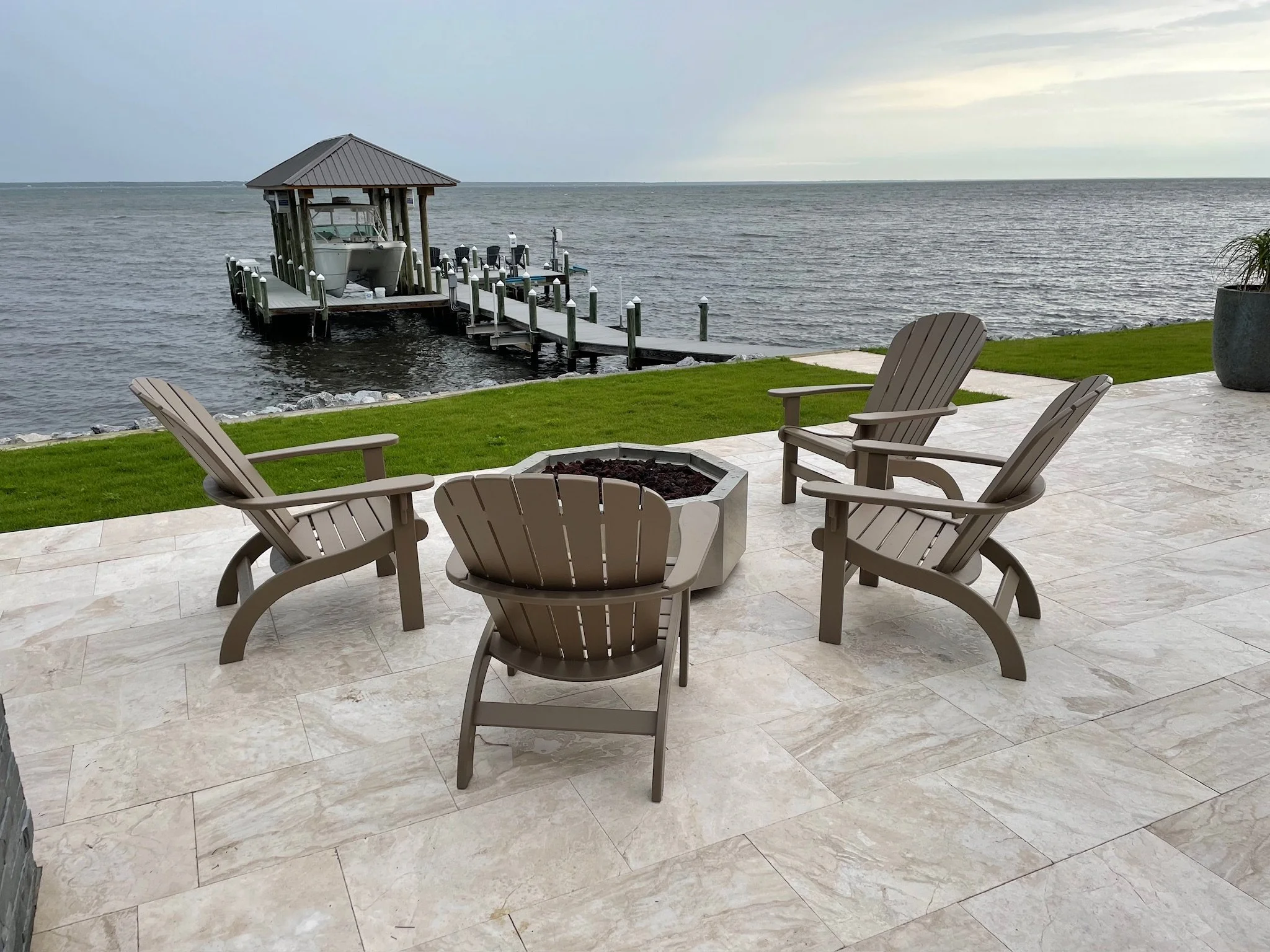Patio with Adirondack chairs around a fire pit overlooking a body of water, with a boat dock, boat, and cloudy sky in the background.
