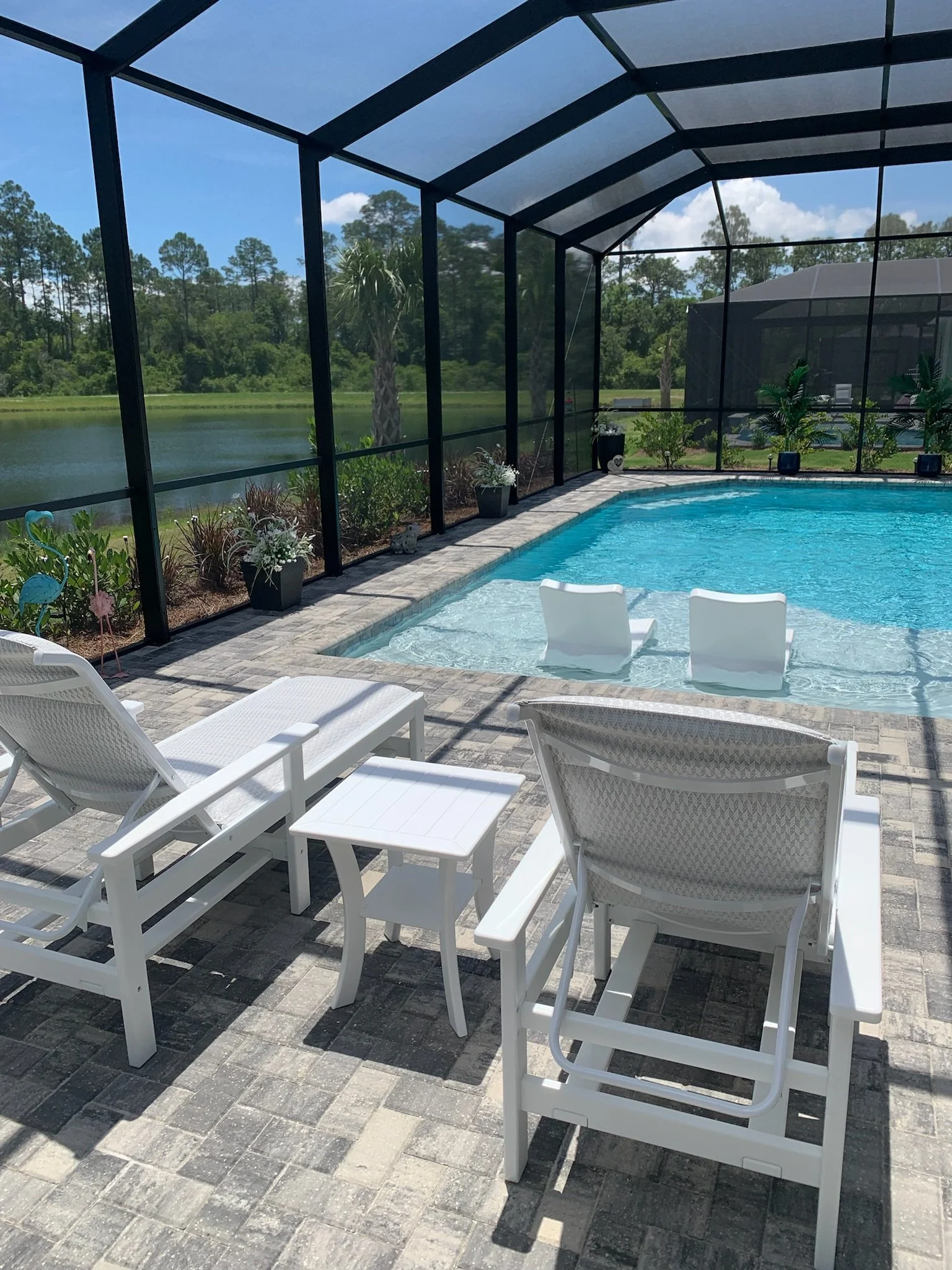 A screened-in backyard pool area with white lounge chairs, a small white table, potted plants, and a view of trees and a lake in the background.