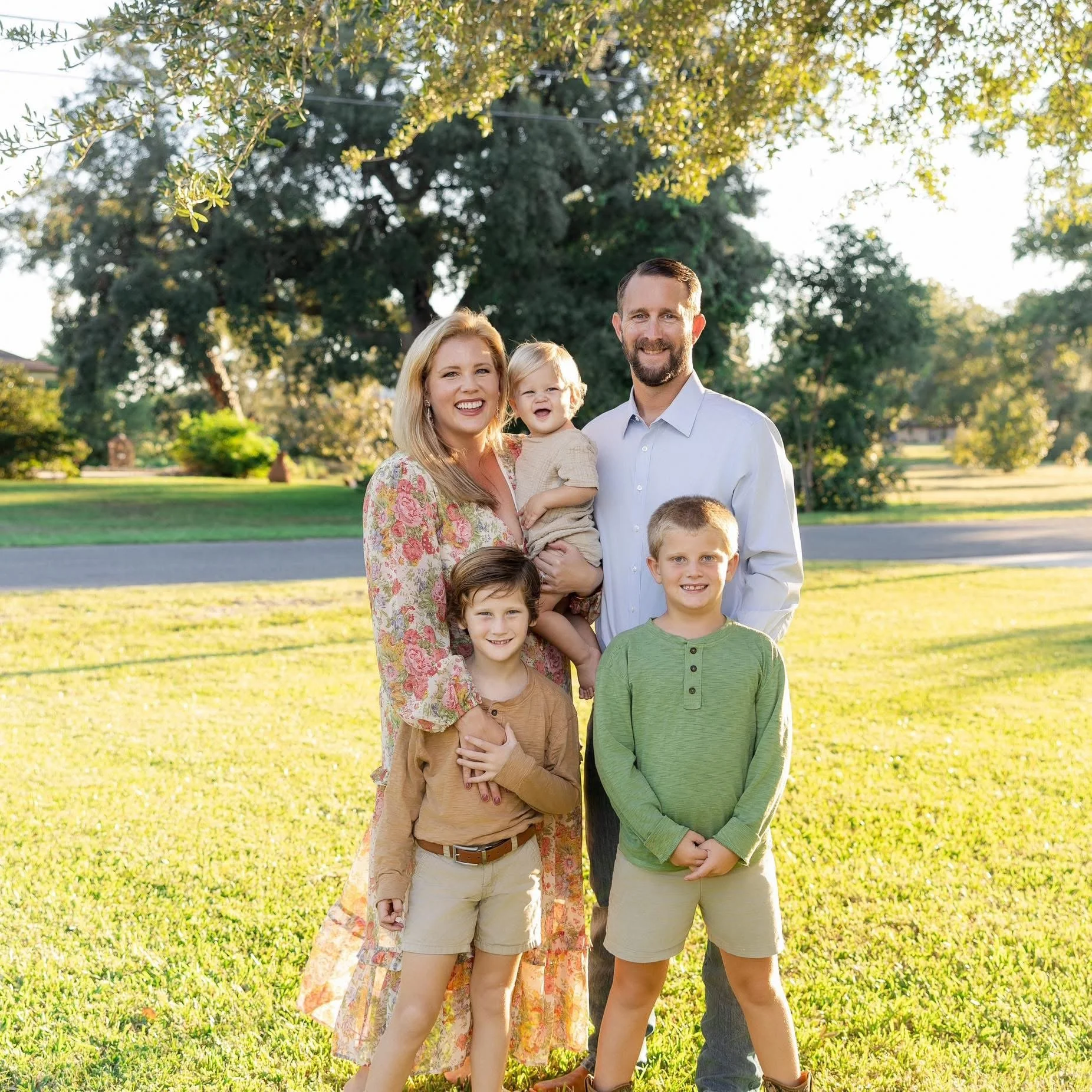 A happy family of six standing outdoors in a park on a sunny day, smiling at the camera. The group consists of two adults and four children, with trees and grass in the background.
