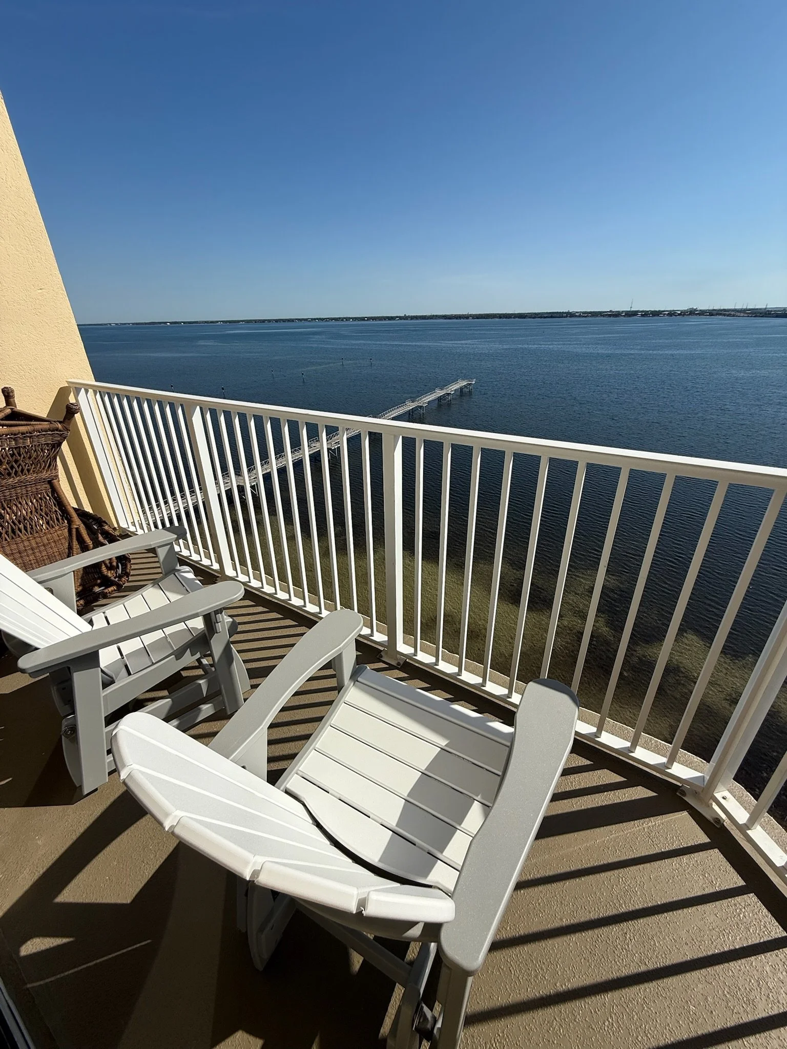 Balcony with white plastic chairs and a railing overlooking a large body of water with a clear blue sky.