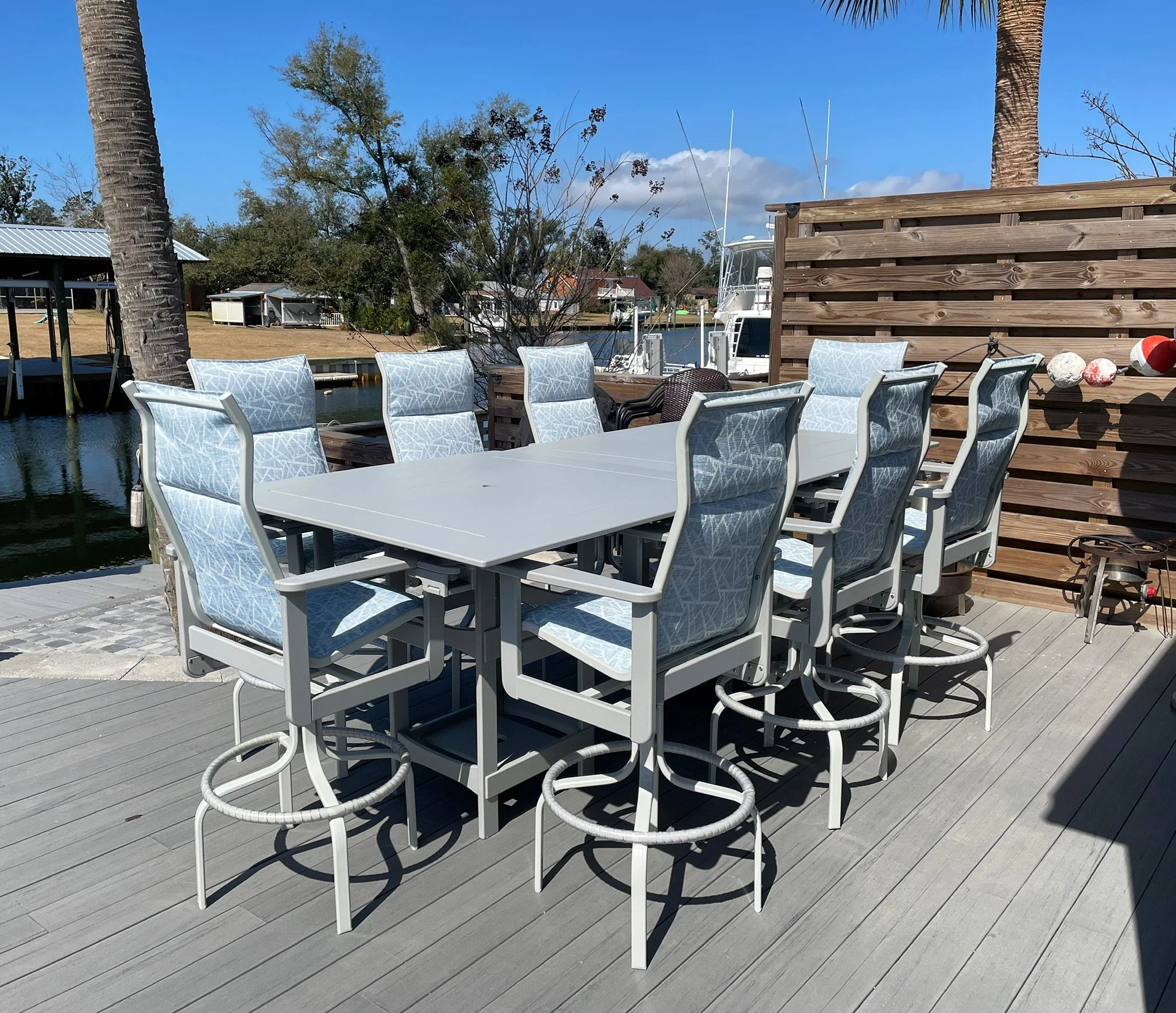 Outdoor patio area with white chairs, wooden privacy screen, palm trees, and a boat docked on the water under a blue sky.