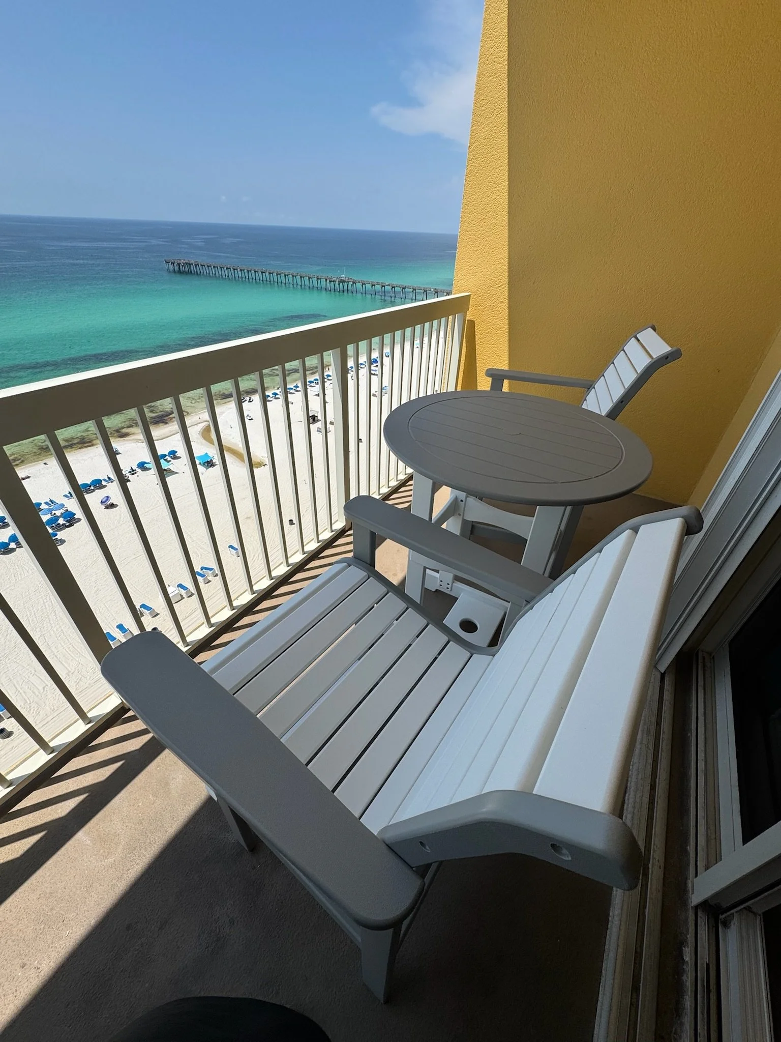 Balcony with two white chairs and a small round table overlooking a beach and ocean with a pier, under a partly cloudy sky.