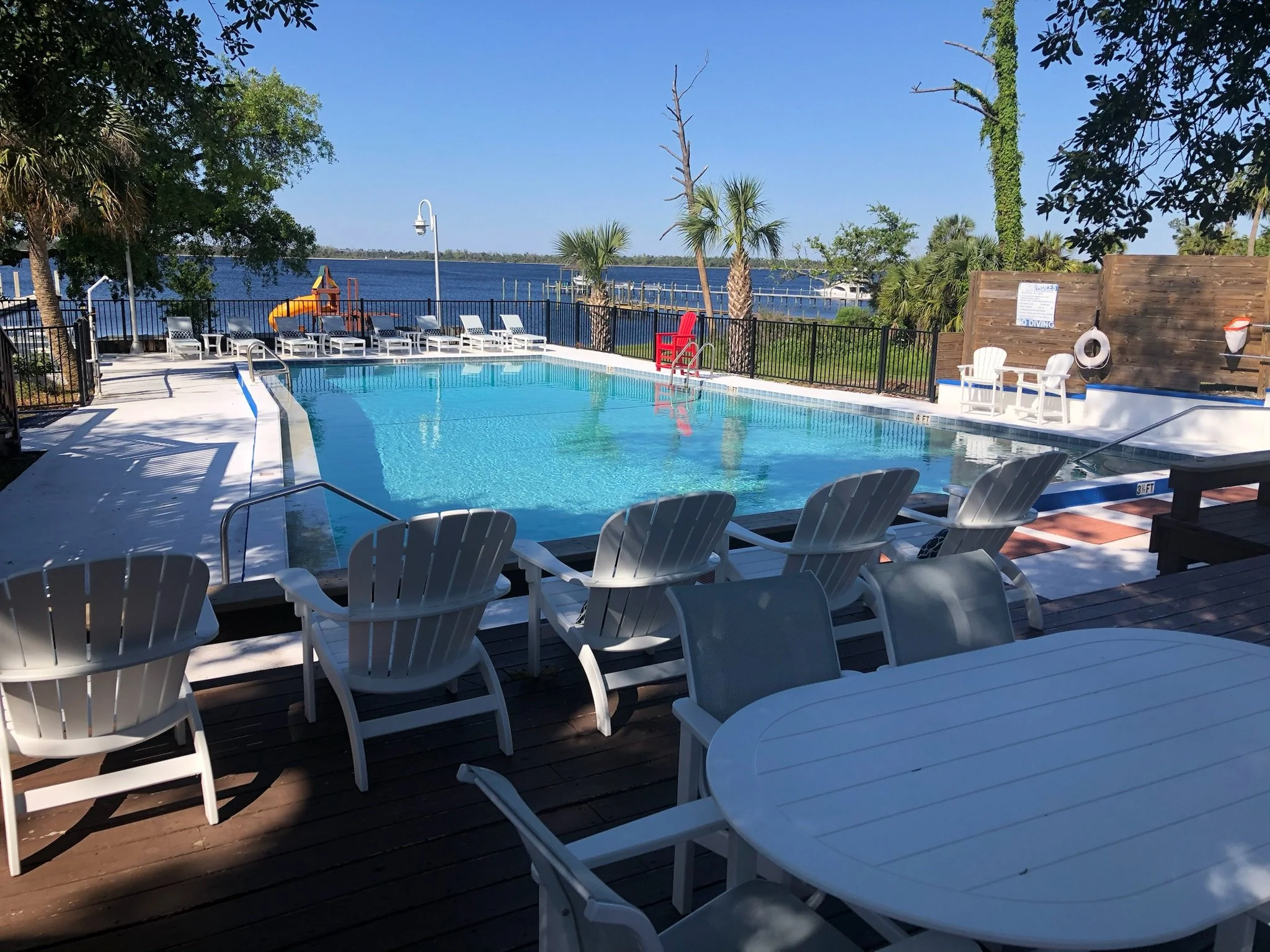 An outdoor swimming pool area with white lounge chairs, some stacked on top of each other, and a slide overlooking a body of water and trees under a clear blue sky.