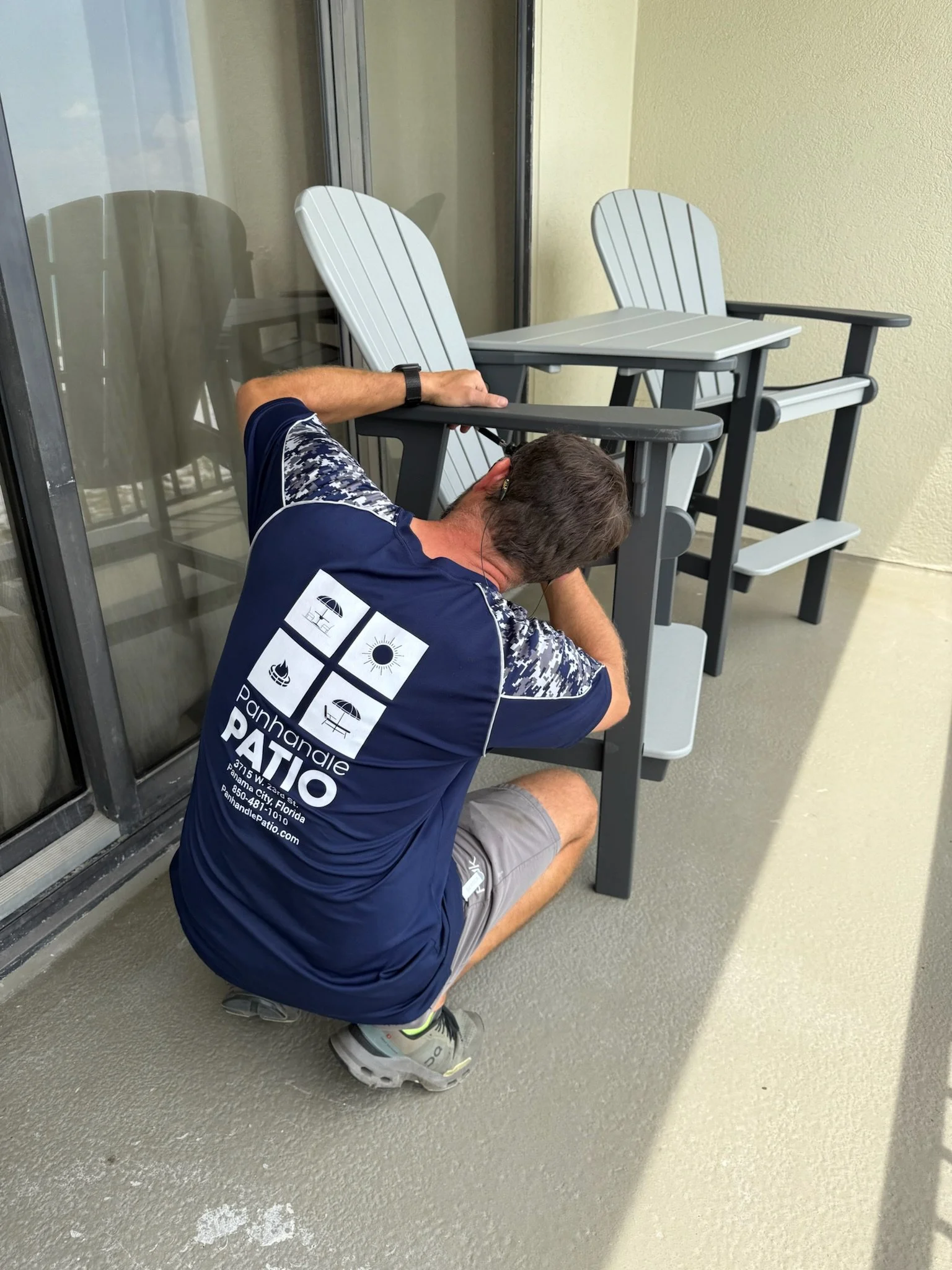 Man kneeling on a balcony, wearing a blue shirt with graphics, gray shorts, running shoes, and earphones, with a collection of outdoor chairs and a small table behind him.