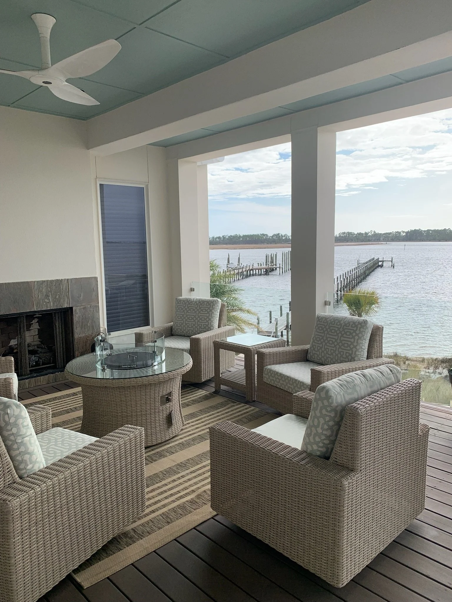Patio with wicker chairs and glass-top table overlooking water and dock with pilings under cloudy sky.