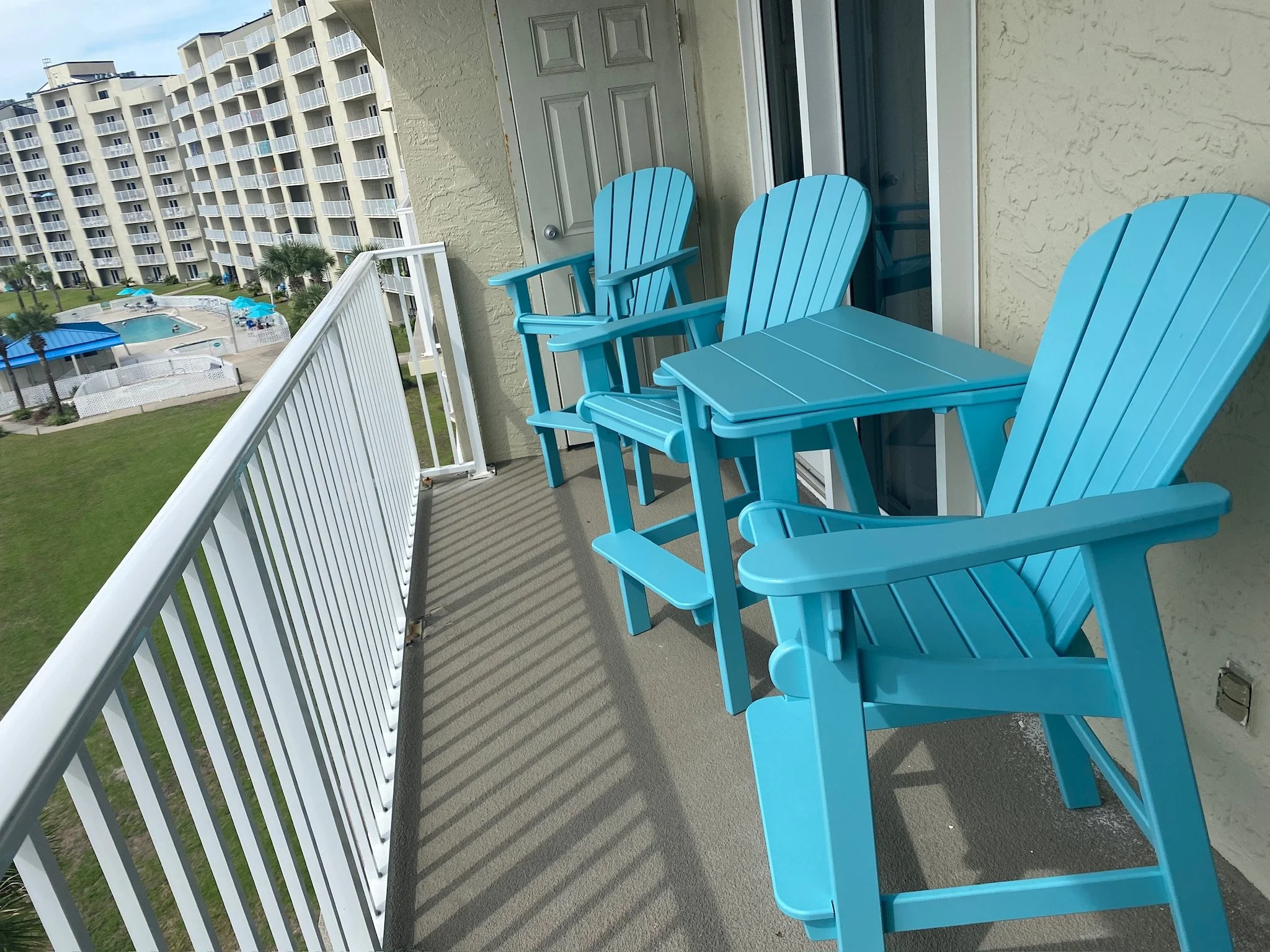 View of a balcony with three blue Adirondack chairs overlooking a resort-style apartment complex and swimming pool area.