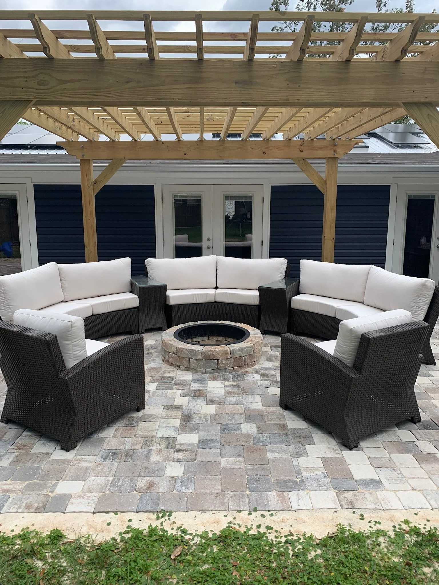 A backyard patio with a fire pit and outdoor seating arranged in a semi-circle on a cobblestone surface. There is a new wooden deck under construction above the patio. The house behind has dark blue siding and white trim, with French doors leading outside.