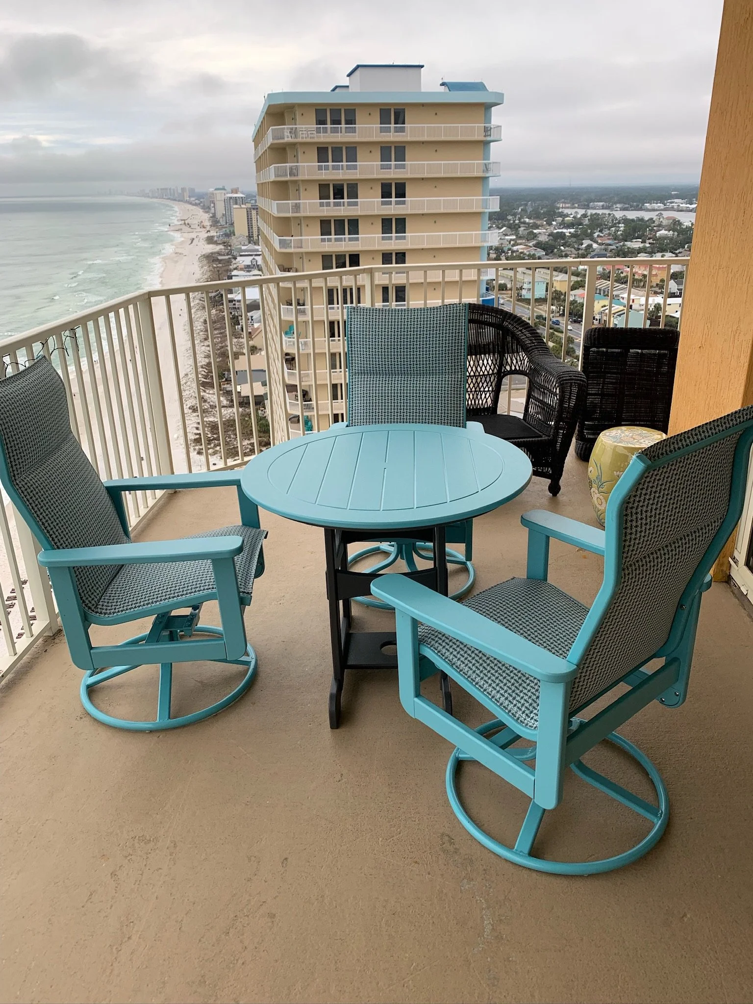 Balcony with blue patio furniture overlooking beach and high-rise buildings, cloudy sky.