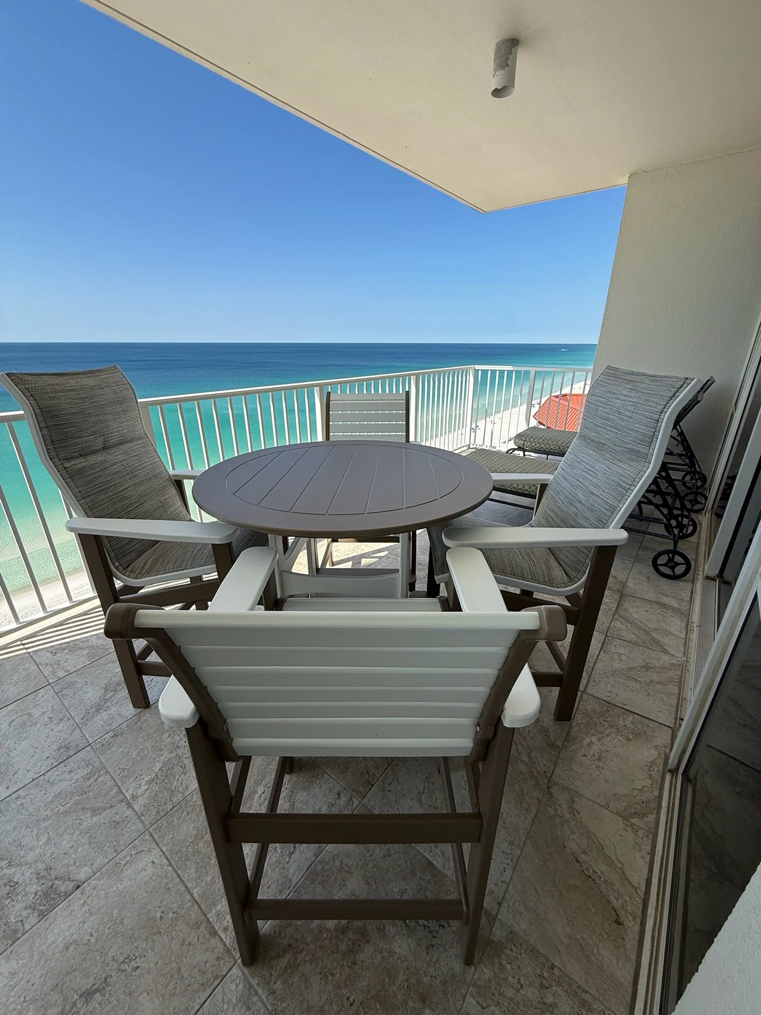 Balcony with a round table and multiple chairs overlooking the ocean with blue sky and beach