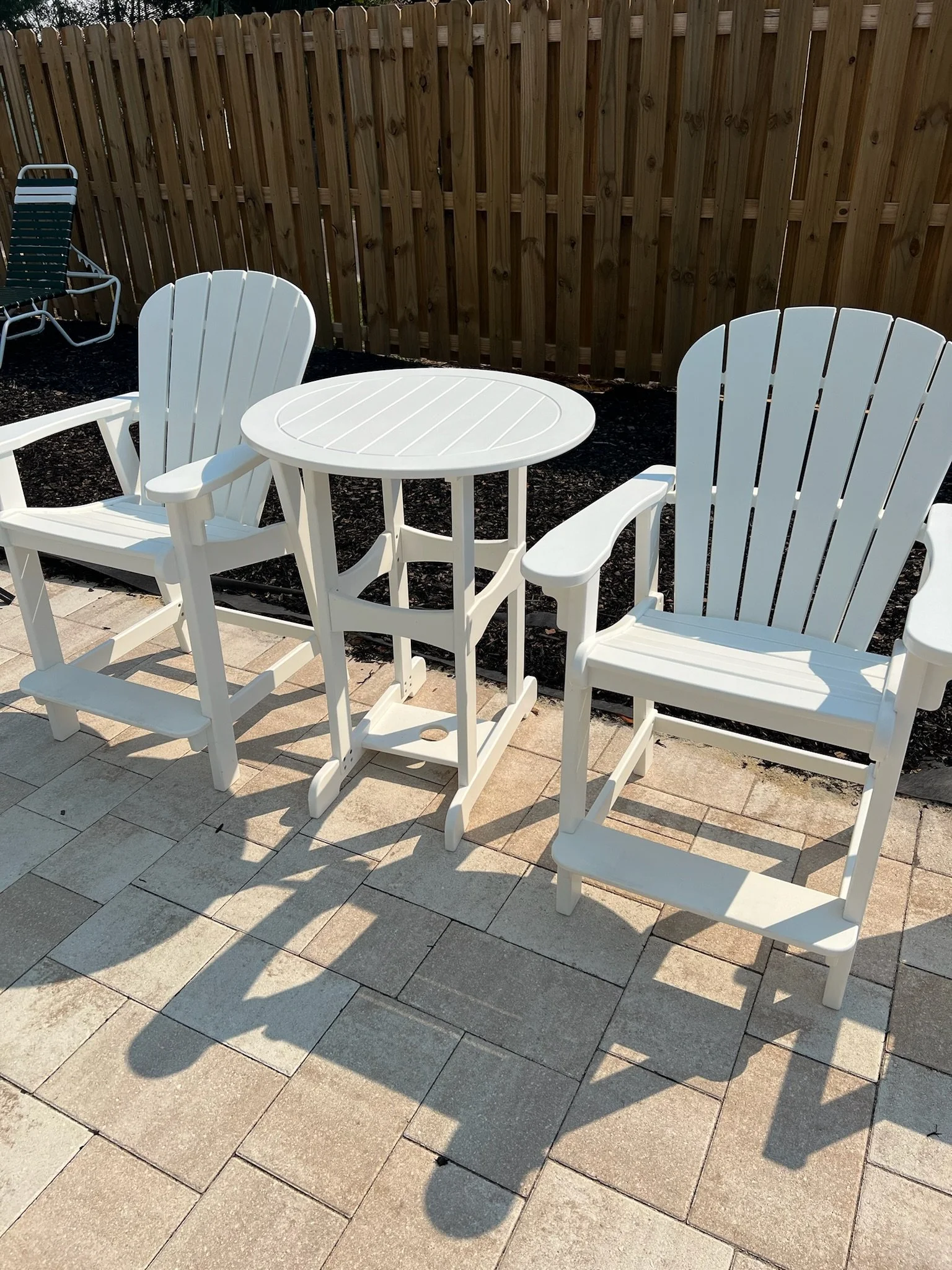 A small outdoor patio area with two white Adirondack chairs and a small white round side table. The patio is paved with light-colored stone tiles and enclosed by a wooden privacy fence. There is also a green folding chair with a white frame placed ag