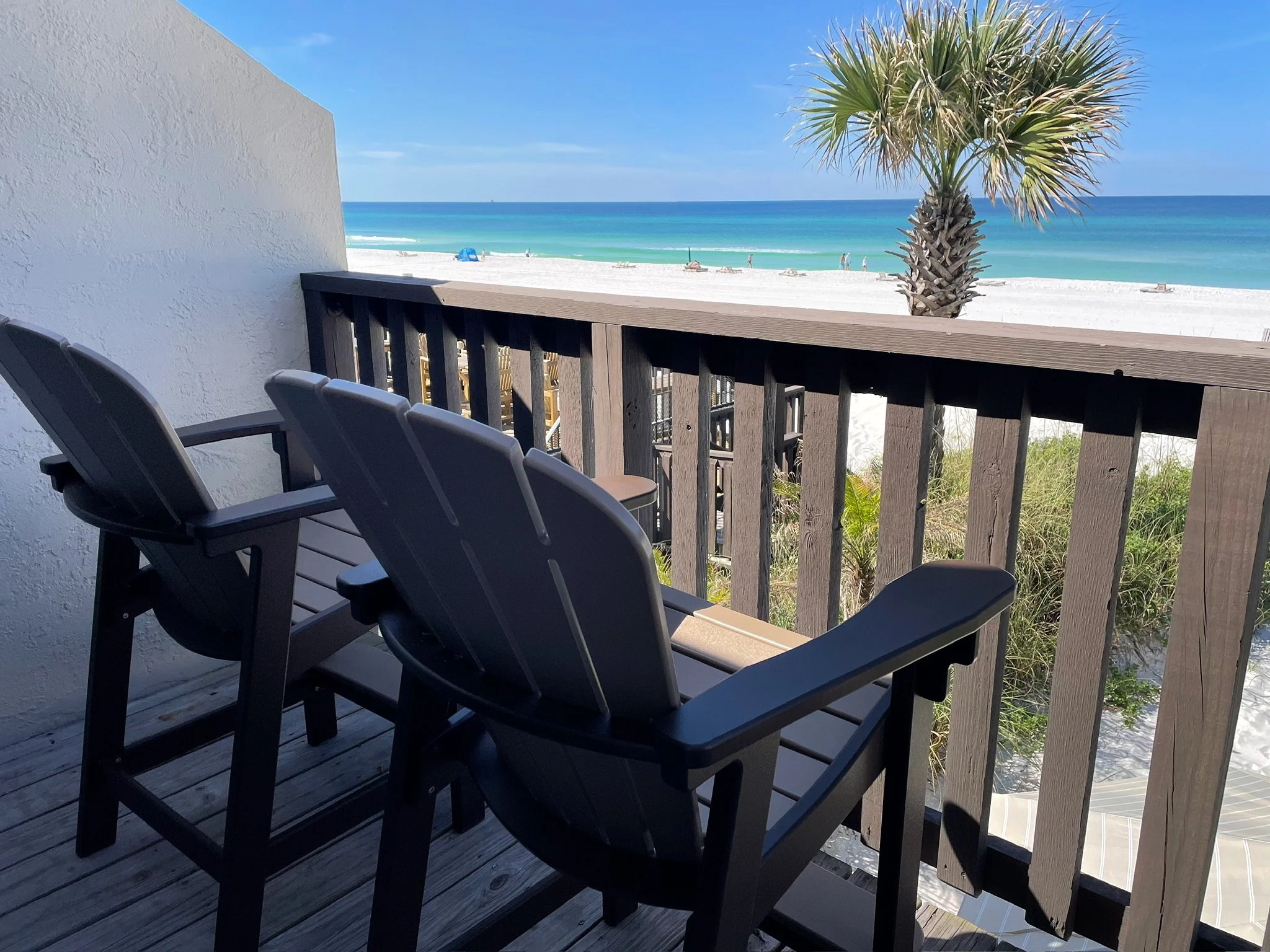 Beach view from a balcony with black chairs and a wooden railing, showing white sand, blue ocean, palm tree, and people walking on the beach under a clear sky.