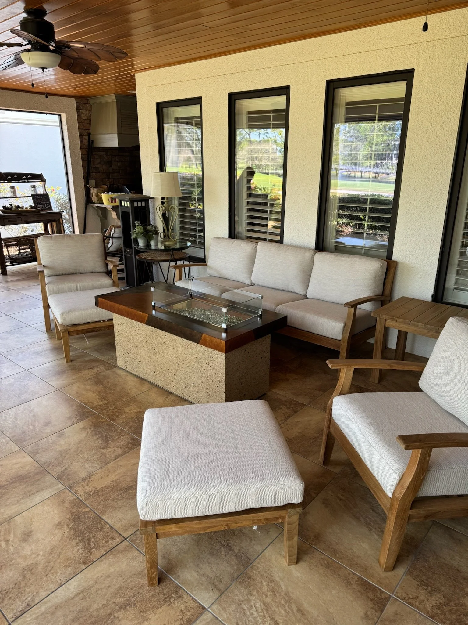 Covered outdoor patio area with beige cushioned chairs, a glass-top coffee table, wooden side table, floor tiles, ceiling fan, and multiple windows reflecting greenery outside.