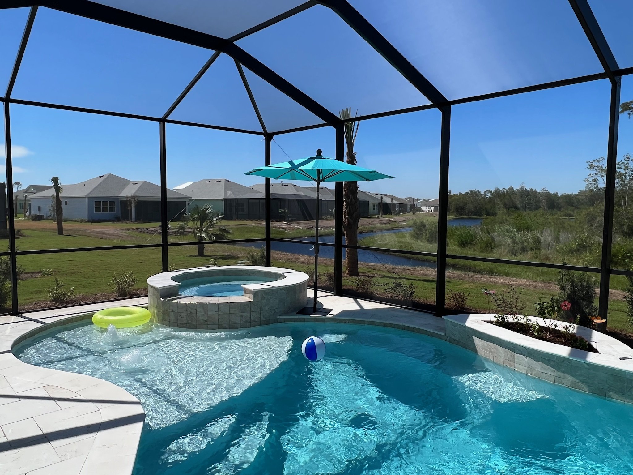 A screened-in backyard with a swimming pool, a blue umbrella, and a hot tub, overlooking a suburban neighborhood with houses, trees, and a small pond under a clear blue sky.