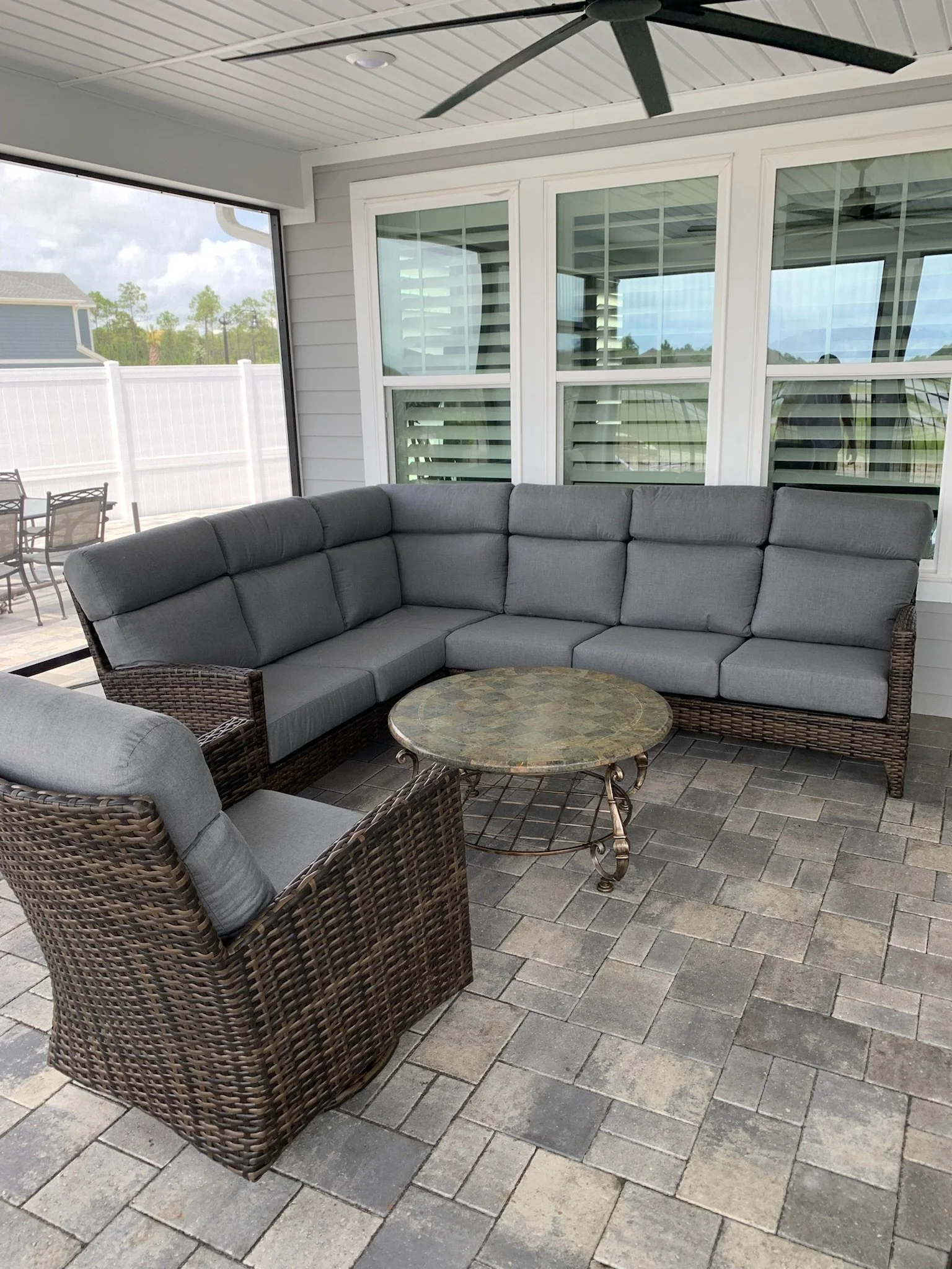 A covered outdoor patio with a ceiling fan, a large sectional sofa with gray cushions, a round coffee table, and a view of a backyard with a patio table and chairs, a white fence, and trees in the background.