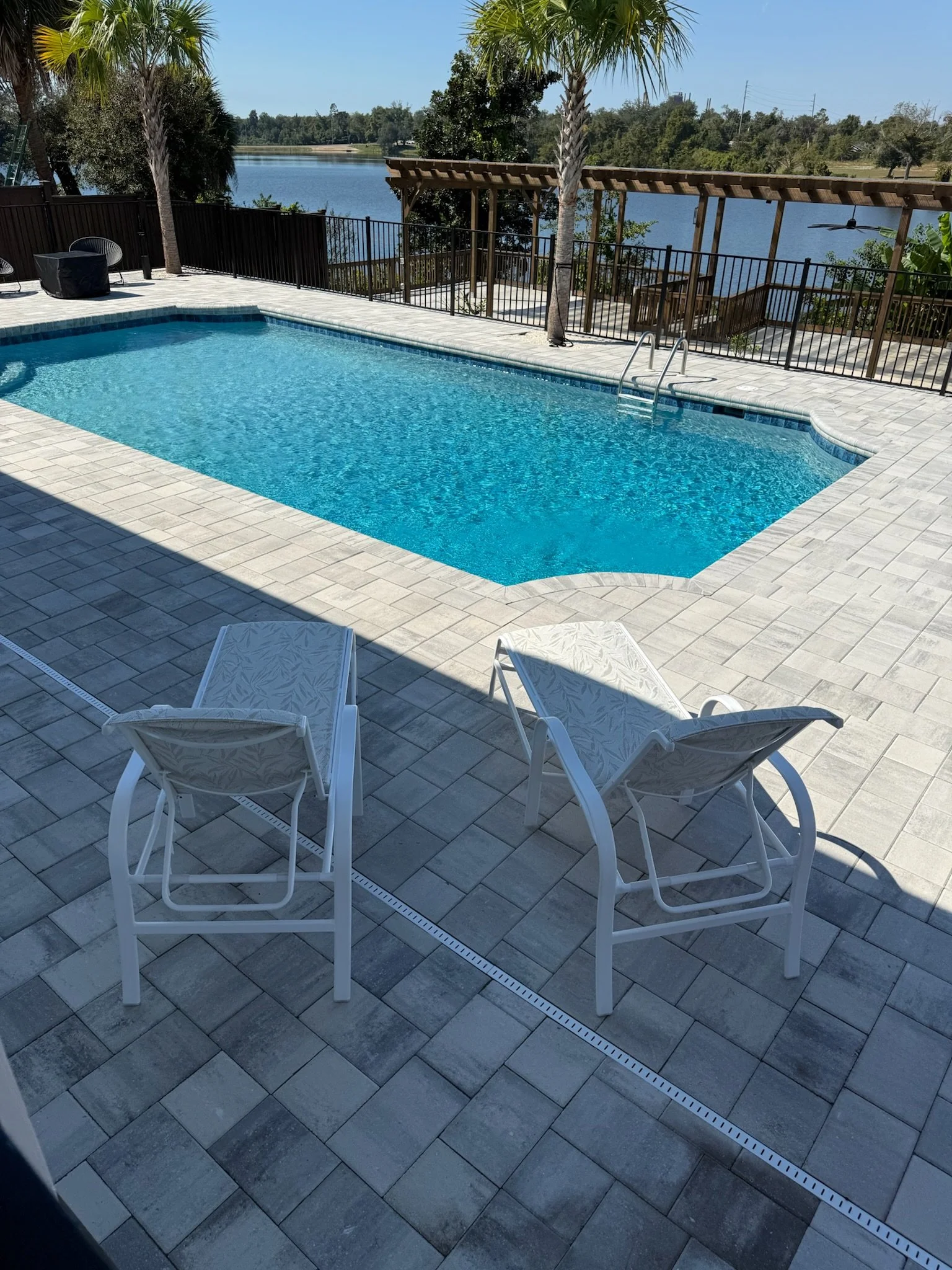 A backyard swimming pool with a tiled deck, surrounded by a black fence. There are patio chairs and a small table near the pool. In the background, there's a lake with trees and a wooden pergola over stairs leading down to the water, with two palm tr