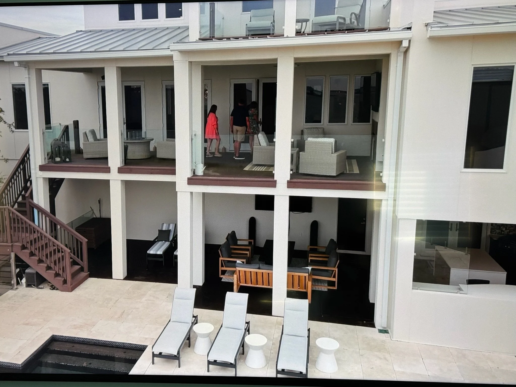 Balcony with outdoor furniture, glass railing, and a staircase leading to the lower level of the house. People are standing near the door.