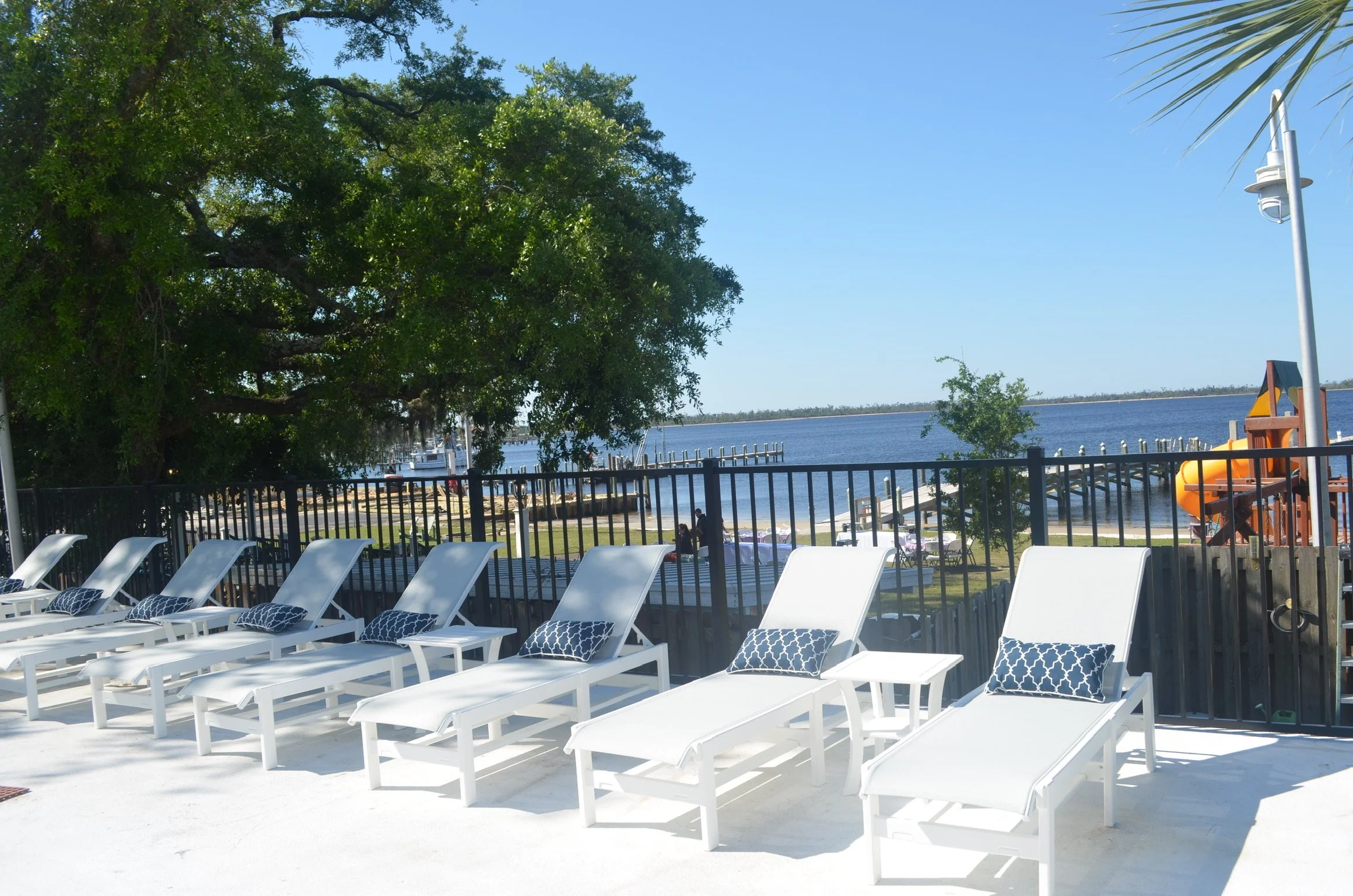 Empty white lounge chairs with blue patterned cushions arranged in rows on a poolside deck overlooking a waterway with boats in the distance. A large leafy tree and a playground slide are visible, with a fence surrounding the area under a clear blue sky.