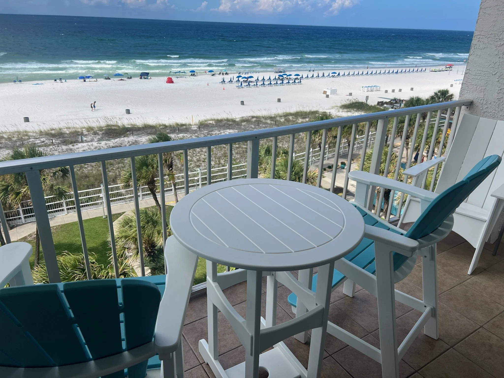 Beach view seen from a balcony with a white outdoor table and chairs, overlooking a sandy beach with blue umbrellas, lounge chairs, and gentle waves in the ocean.