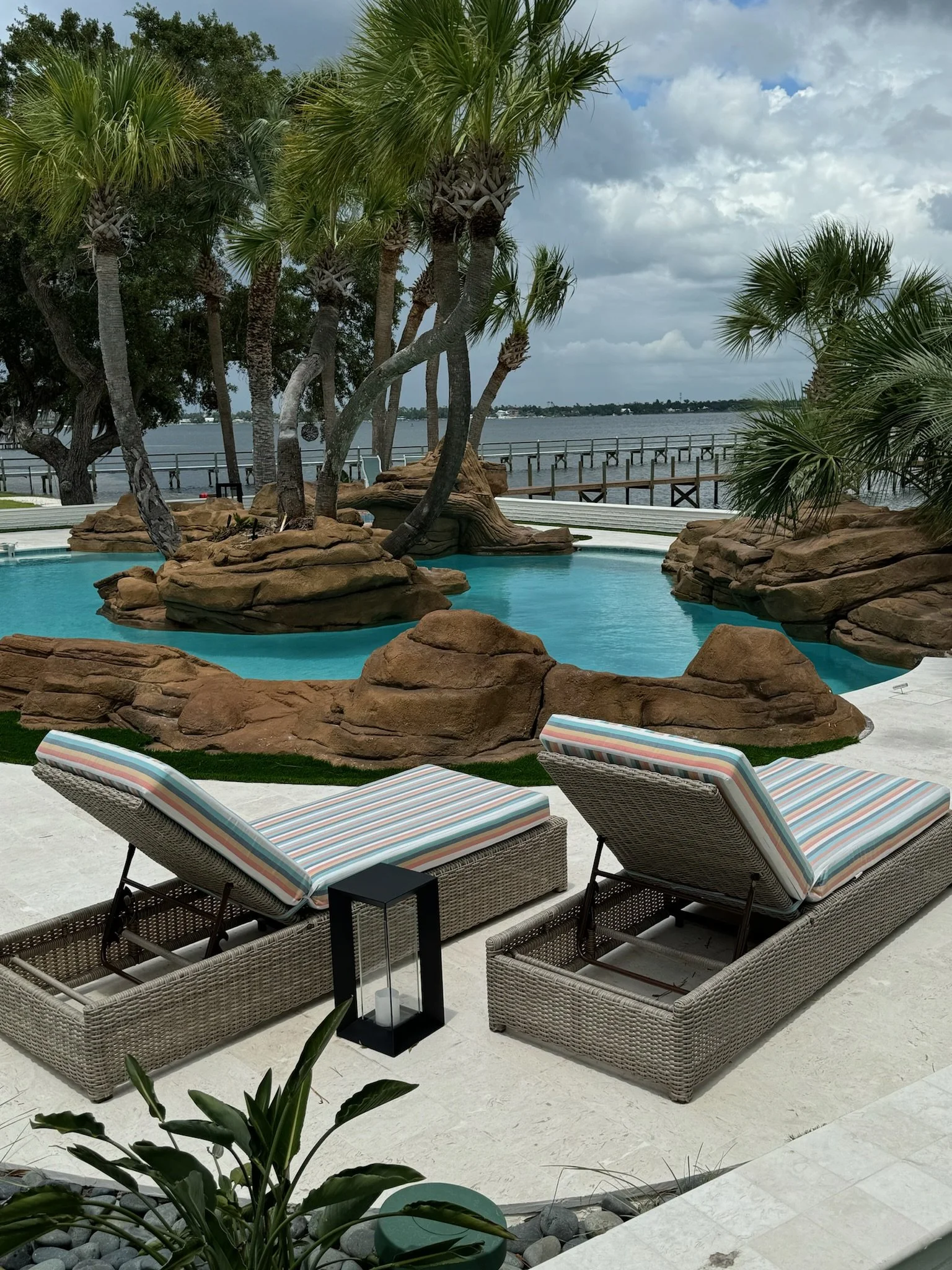 Pool with rock surroundings and palm trees near water, lounge chairs with striped cushions, viewed from a patio with plants and a railing.