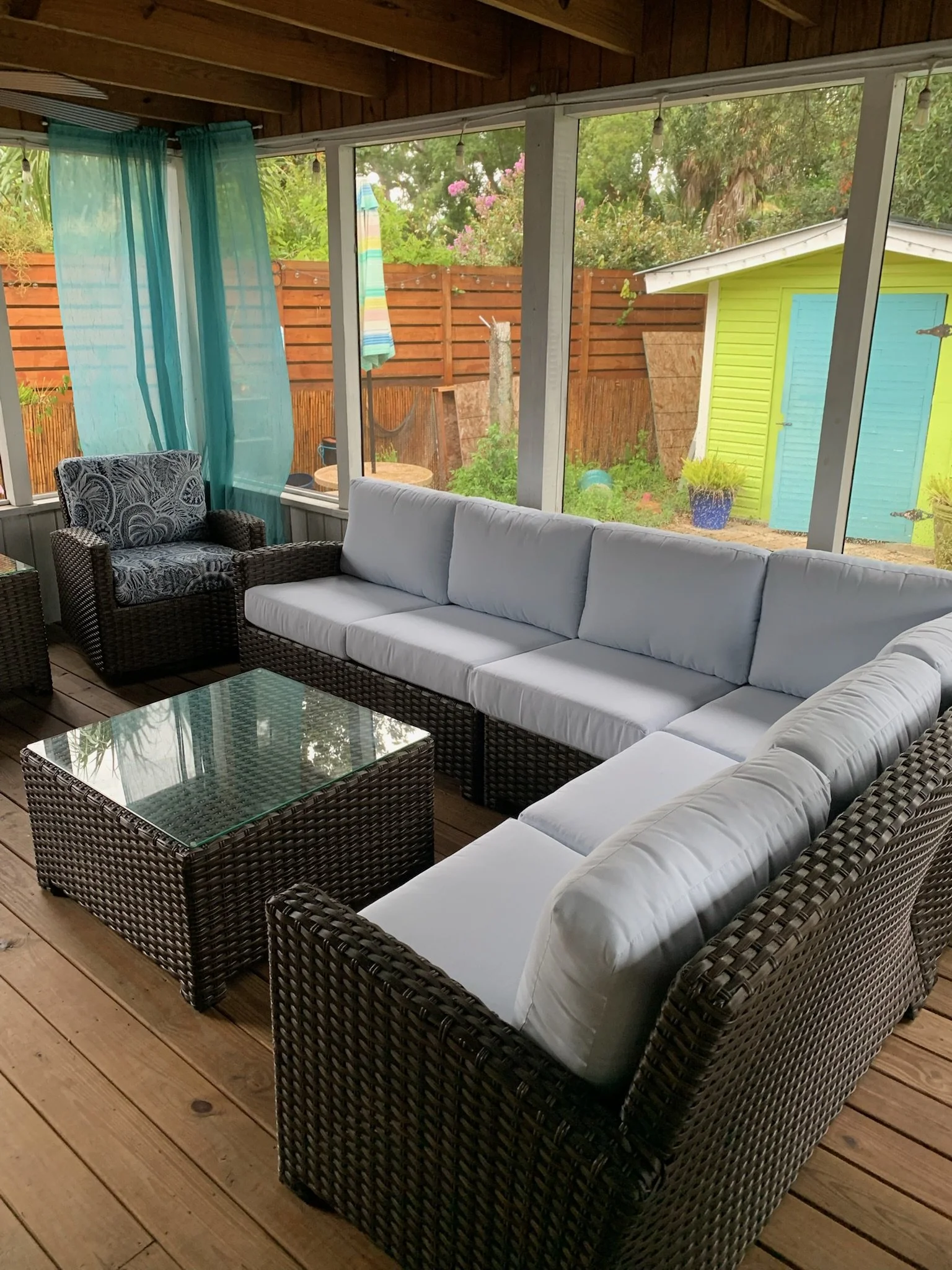 A wicker patio sofa with white cushions, a matching wicker chair with patterned fabric, a glass-topped wicker coffee table, inside a screened porch with wooden floors and a wooden ceiling, overlooking greenery and a small yellow shed outside.