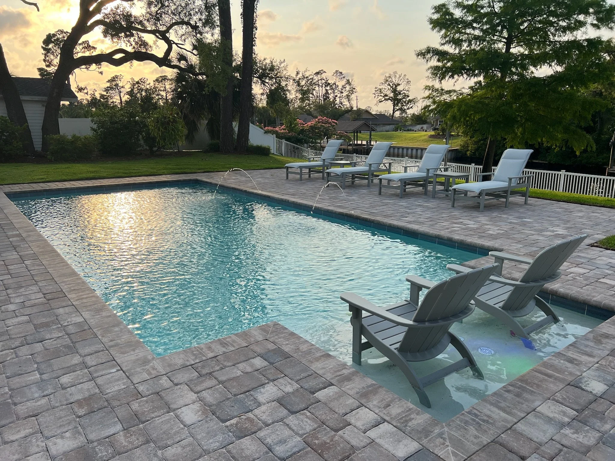 A backyard with a swimming pool and four lounge chairs on a brick patio, surrounded by trees, plants, and a white fence, during sunset.
