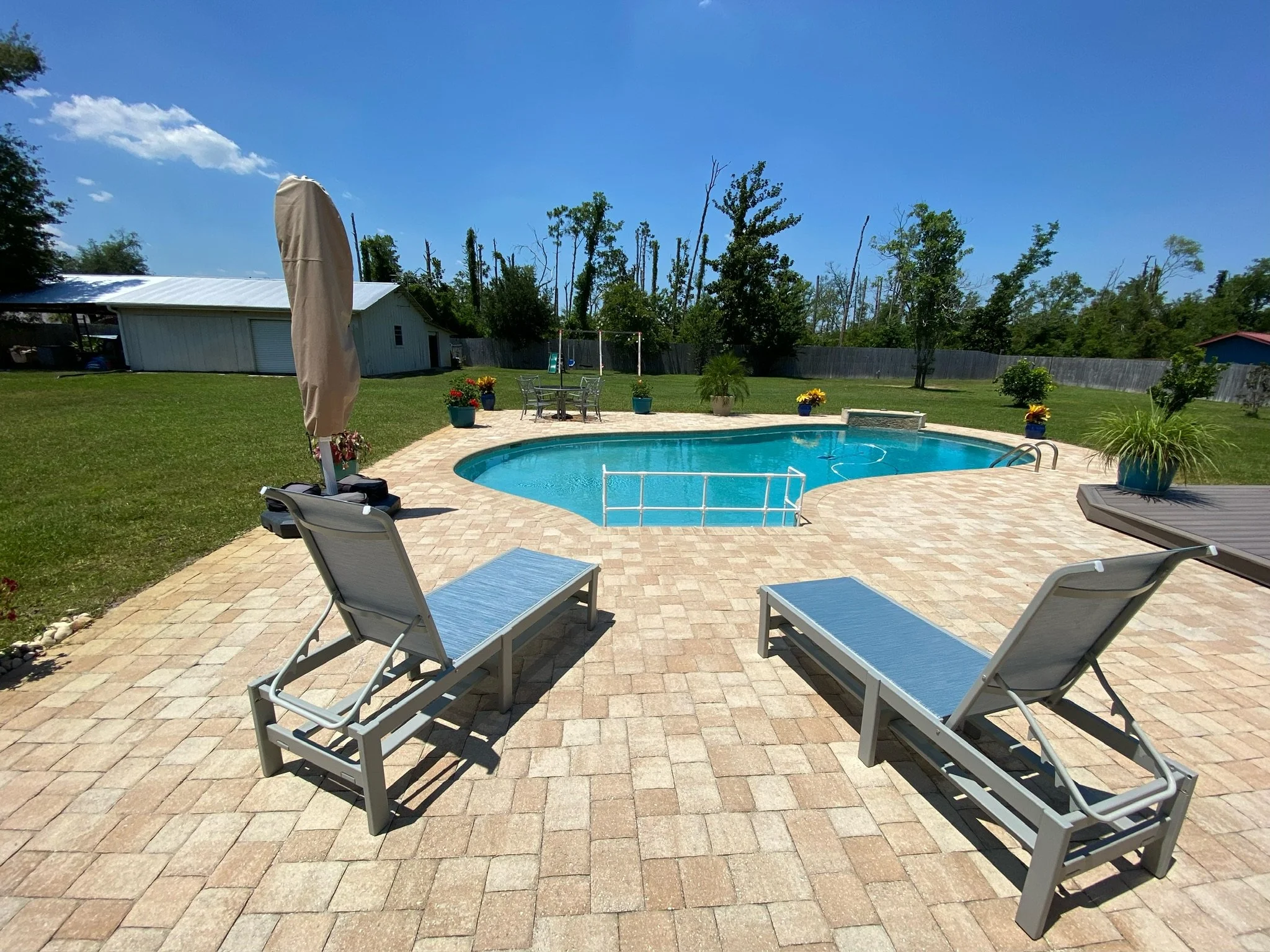 A backyard with a swimming pool surrounded by a brick patio, outdoor furniture, and potted plants. There is a white shed, a swing set, and a tall wooden fence with trees in the background under a clear blue sky.