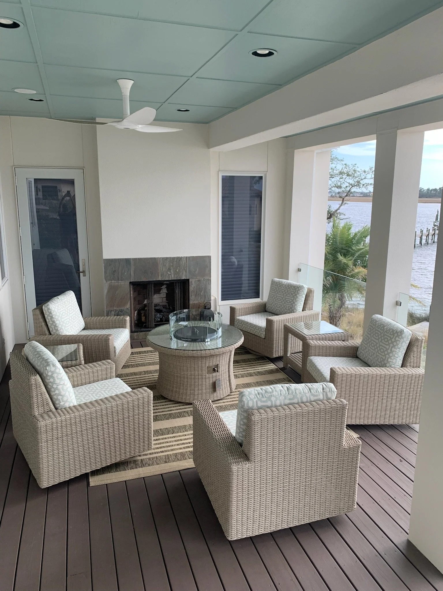 Outdoor patio area with beige wicker chairs, glass-topped table, fireplace, and a view of water and trees outside.