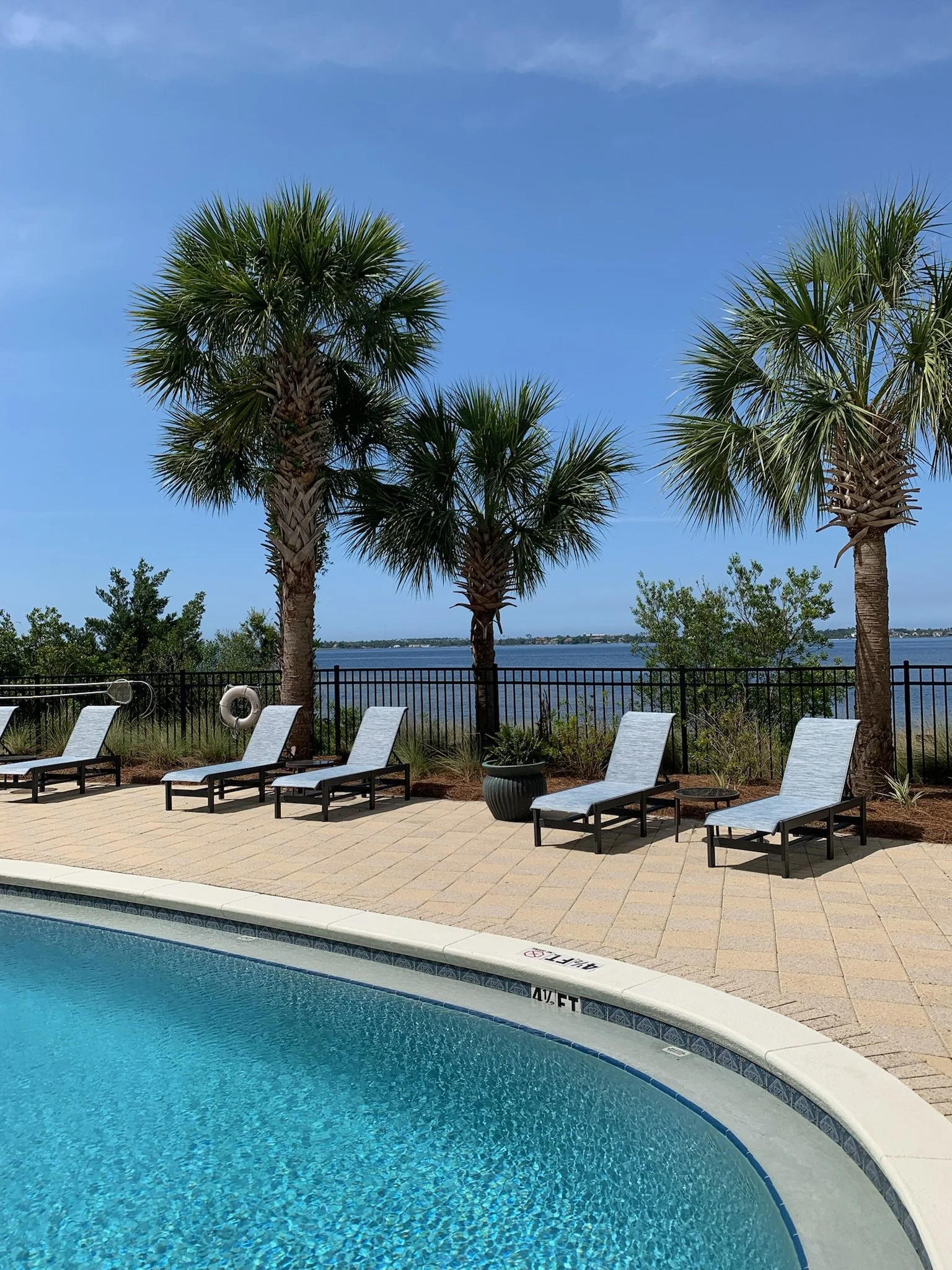 A swimming pool with blue water, lounge chairs on a patio, palm trees, and a body of water in the background under a partly cloudy sky.